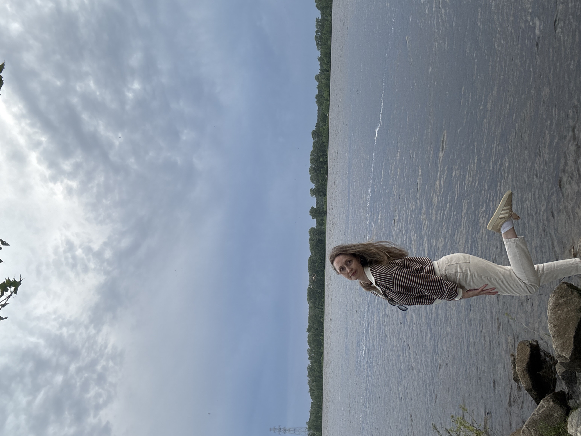 A woman stands on a rocky shore next to a body of water, possibly a lake or river. The background shows a line of trees across the water, with a pale, cloudy sky above. An electrical power tower is visible in the distance. The woman is wearing cream-colored trousers, a striped shirt, and off-white sneakers. The setting appears to be a natural area, possibly a park or nature preserve near the city of Chaikovsky in Russia.