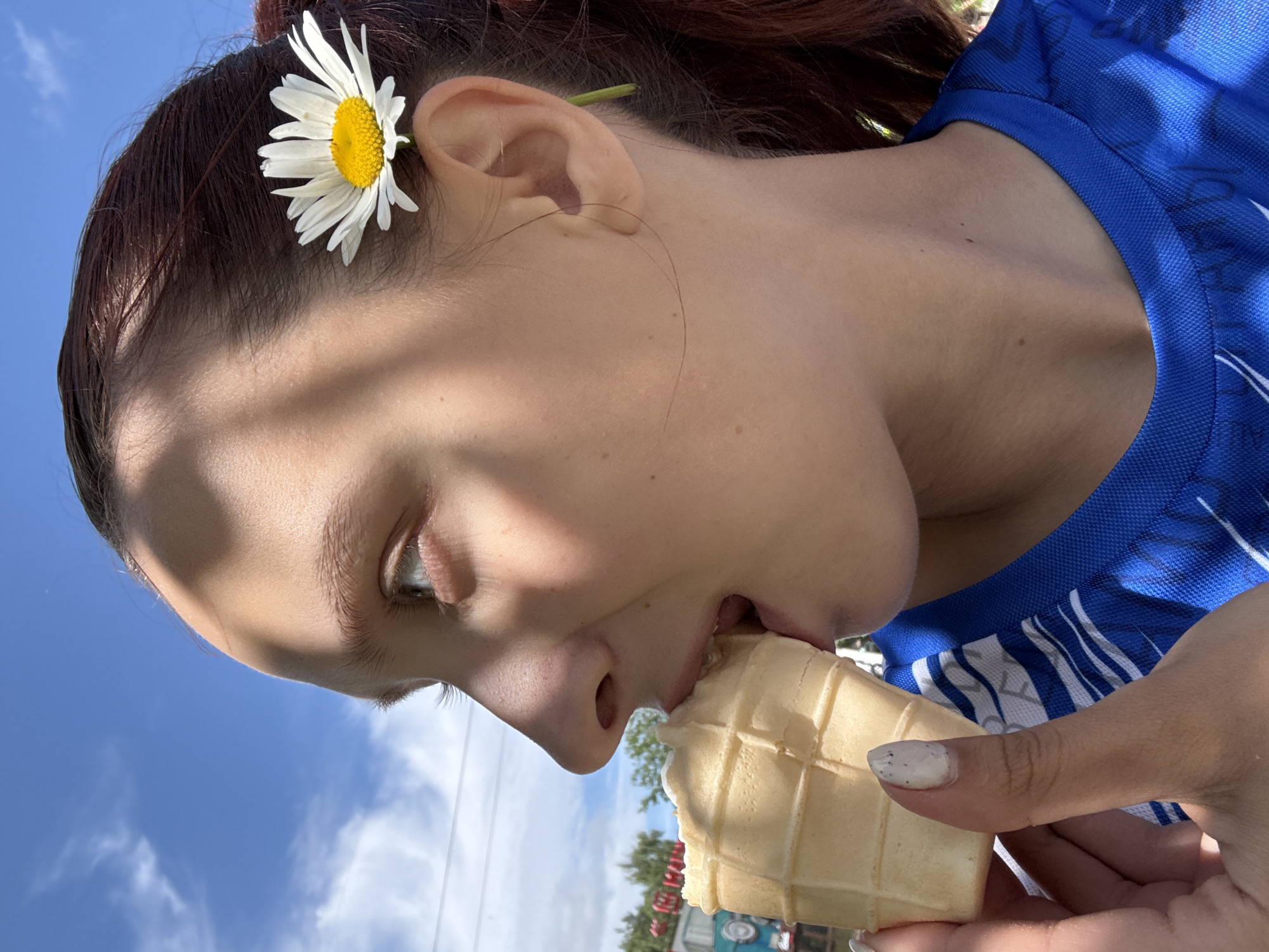 A profile close-up shows a young woman with brown hair, a daisy tucked behind her ear, eating an ice cream cone. She is wearing a blue t-shirt. She is outside under a bright blue sky with white clouds. The background is blurred, but appears to show trees and some structures, suggesting an outdoor public space like a park or fairground. The lighting suggests it is daytime. The image does not contain enough information to determine the specific location in Chaikovsky, Russia.