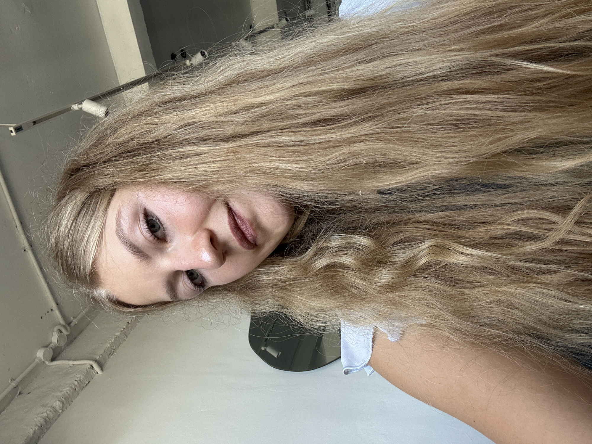 A close-up, tilted-angle photograph shows a fair-skinned young woman with long, light blonde hair. Her head is positioned to the left. Her eyes are open and gazing toward the camera. Her lips are closed. A white shirt sleeve is visible on the right side of the frame. In the background, are electrical cables, concrete wall and a black, round object. The image was not taken in Kuta Selatan, Indonesia.