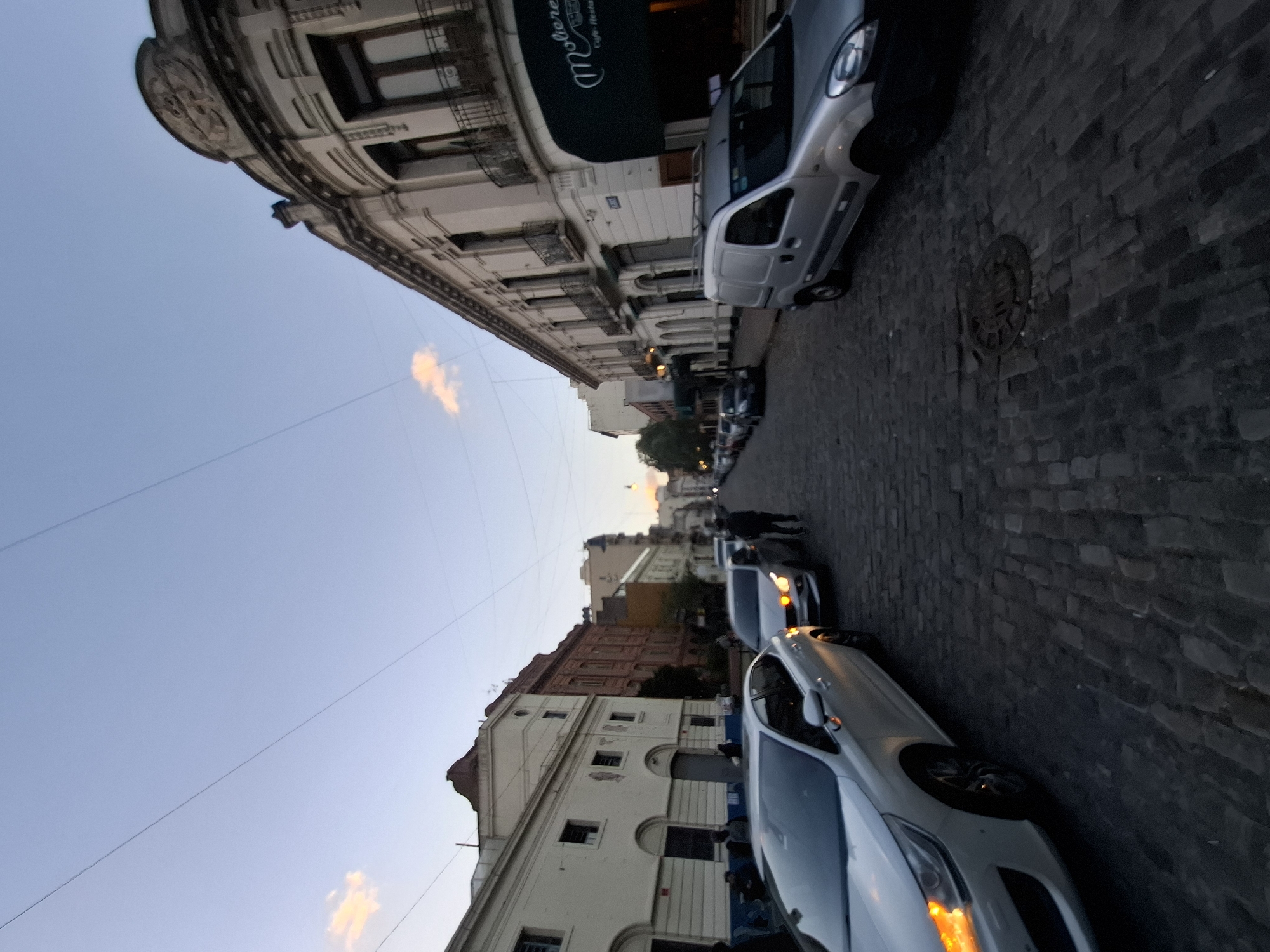 A street scene in Buenos Aires, Argentina. The cobblestone street is lined with parked cars on both sides. Buildings with intricate facades flank the street, and a cafe awning is visible on one side. The sky is blue with some light clouds. Pedestrians can be seen walking in the distance.