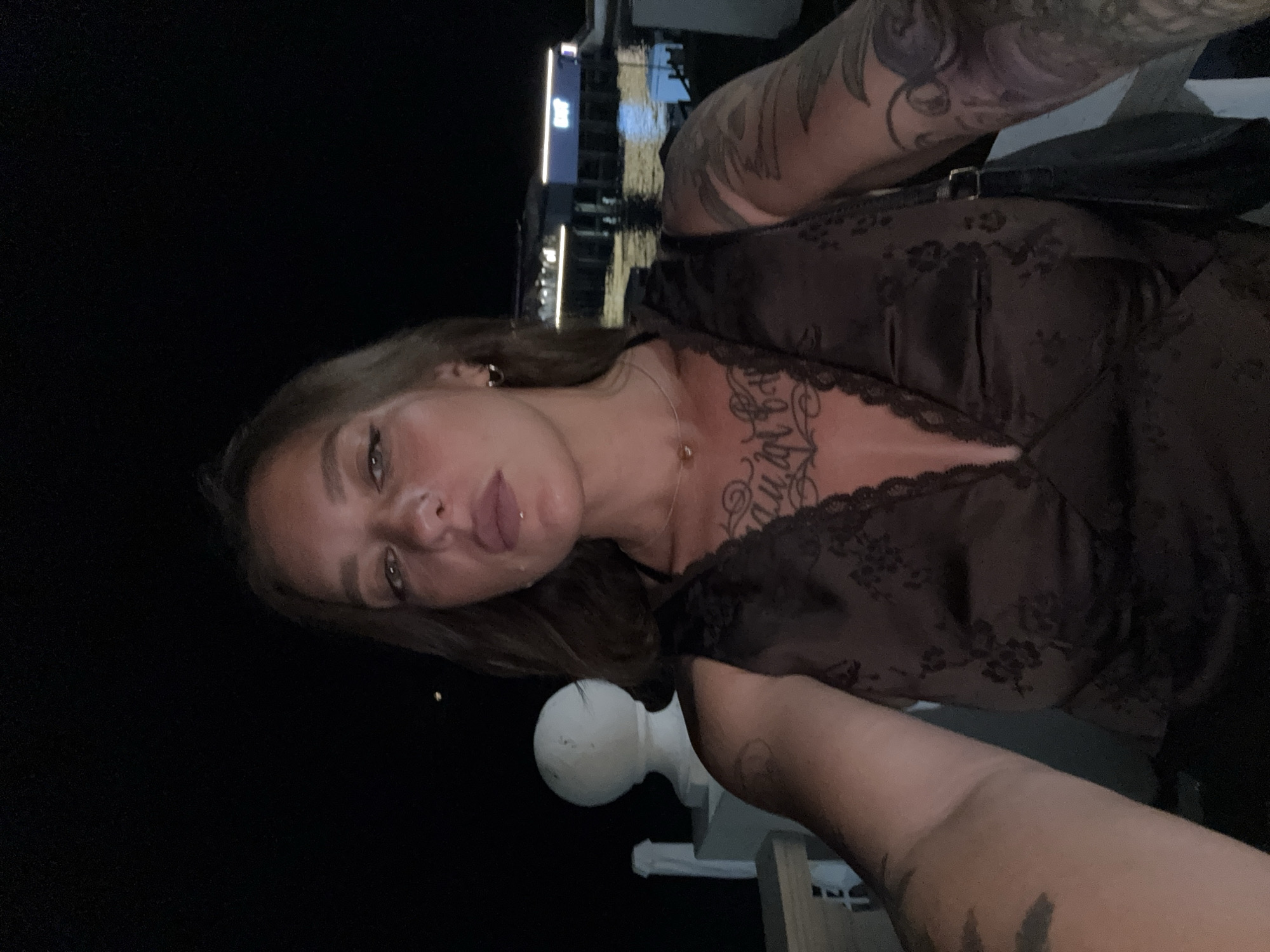 A woman is taking a selfie at night on a balcony overlooking a body of water. She has tattoos on her arms and chest and is wearing a dark brown, lacy top. In the background, there is a railing, a white post with a spherical top, and a building with illuminated signs across the water. The overall impression is that the photo was taken in Sochi, Russia, potentially near the coast or a riverfront area, given the presence of water and the architecture suggestive of a resort town.