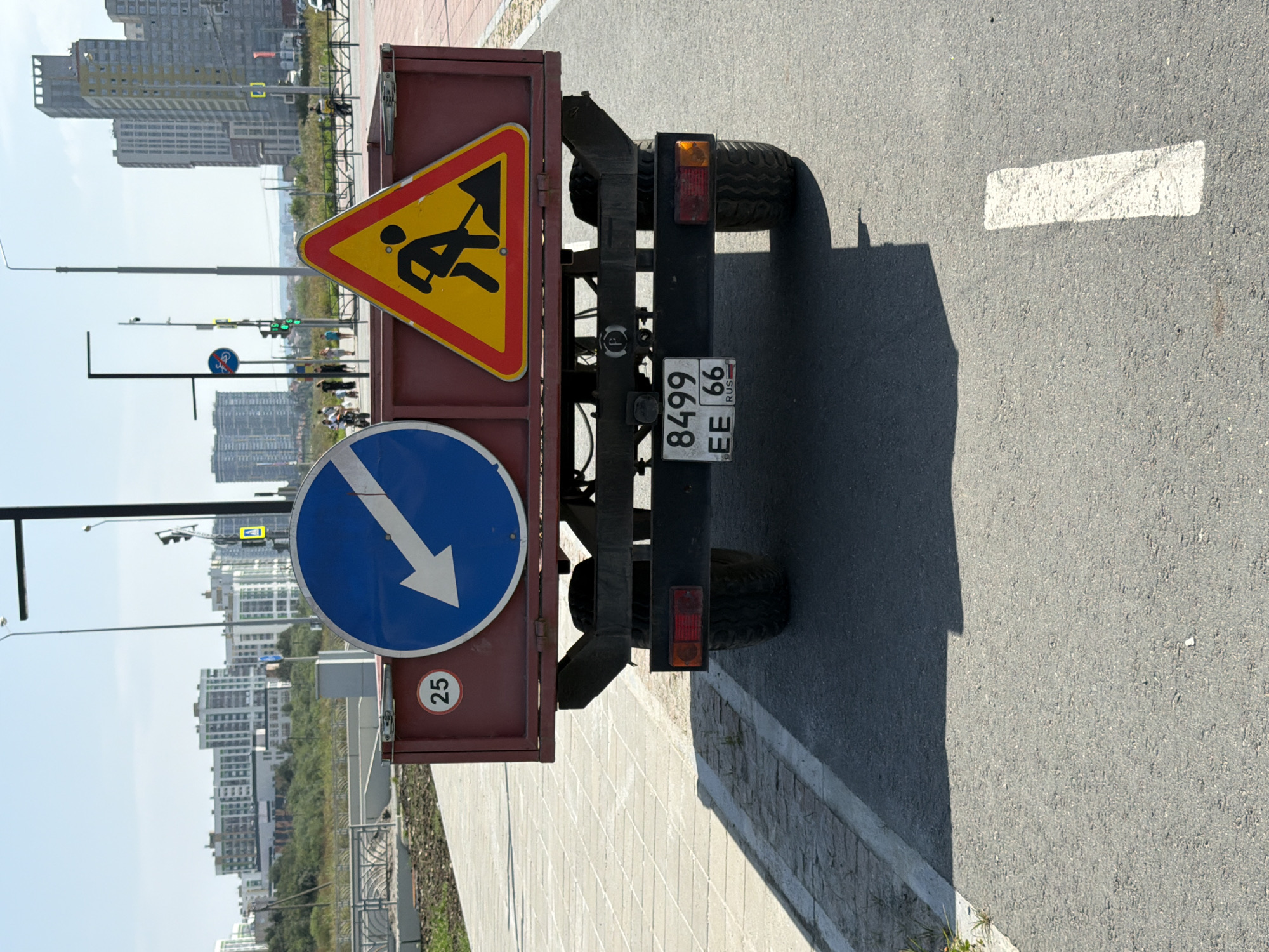 The image shows the rear of a trailer on a paved road in Yekaterinburg, Russia. The trailer is red and has two signs attached to its side. The left sign is a blue circle with a white arrow pointing down and to the left. Inside the circle, on the bottom left, is a white circle with the number "25" in black. The right sign is a yellow triangle with a red border and a black silhouette of a person digging with a shovel. A license plate on the rear of the trailer reads "8499 EE 66 RUS". In the background, there are several modern buildings and a clear sky. The road appears to be a bike path or a pedestrian walkway with a concrete border on the left.