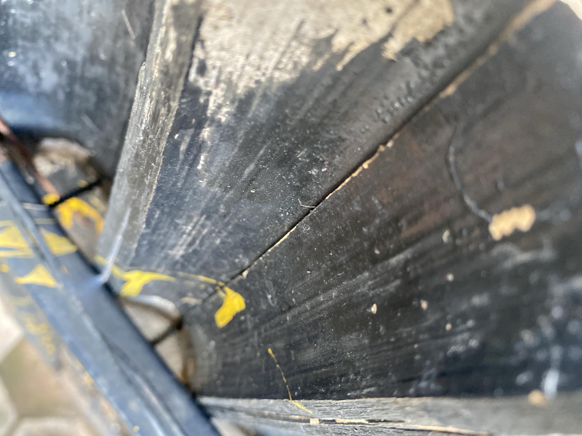 The image shows a close-up, angled view of weathered, black-painted wooden stairs. Yellow paint splatters and streaks are visible on the stairs, suggesting they may have been marked or decorated. The wood appears worn and aged, with visible grain and some splintering. The background is out of focus but suggests an outdoor environment. There are no people or other distinct objects visible in the frame. The specific location in Gura Top, Nigeria cannot be determined from the visual information provided.
