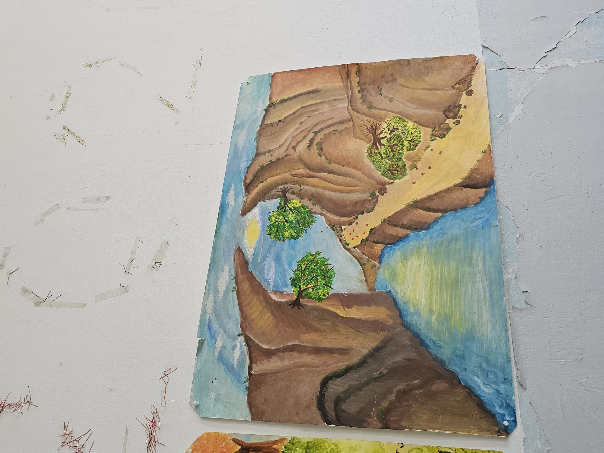 A painting is hanging on a white wall with peeling paint. The painting depicts a canyon landscape with brown cliffs, green trees, and a blue river or lake. There is also some tape and tinsel stuck to the wall. The scene is indoors. There is no information about the city of Yerevan, Armenia.