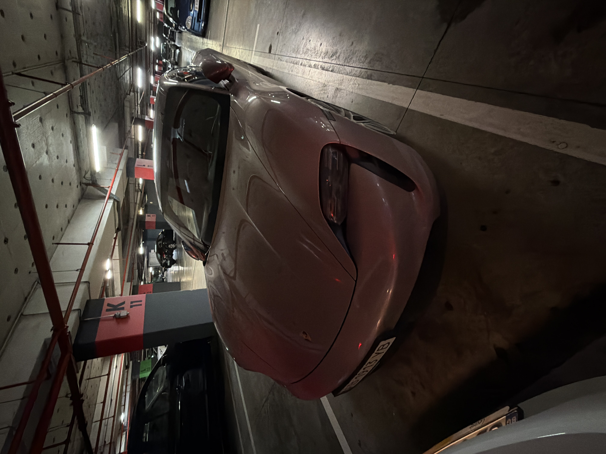 The image depicts a pink Porsche Taycan parked in an underground parking garage. The location is Burgas, Bulgaria, based on the license plate number. The car is angled slightly to the right and faces forward. The garage is dimly lit, and other parked cars are visible in the background. The garage walls are concrete, and there are red pipes and parking zone markers visible.