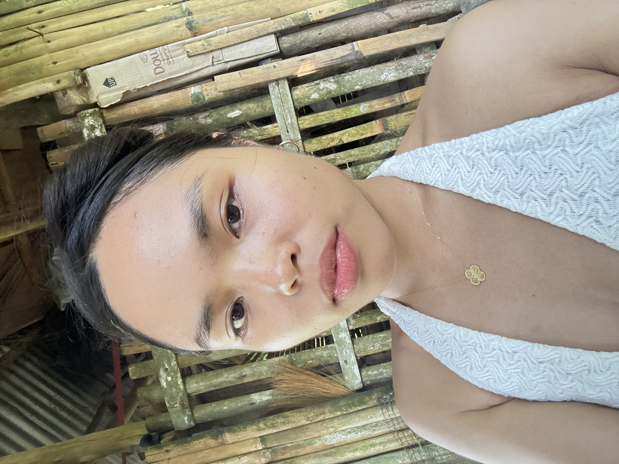A young woman is depicted in a direct, close-up, selfie-style portrait, positioned outdoors. She has dark hair pulled back, dark brown eyes, and full lips with a light pink gloss. She wears a white textured halter-neck top and a gold-colored necklace with a four-leaf clover pendant. A small gold hoop earring is visible in her right ear. The background consists of a rustic, vertical bamboo and horizontal wooden slat structure, suggesting a fence or wall. Many bamboo poles exhibit green moss or algae growth. In the upper left, a corrugated metal roof is partially visible. To the left of the woman, a bundle of dried fibrous material, resembling a broom, is attached to a pole. On the right, a section of a cardboard box with an "UP" arrow and partial text "Dow" (likely "Downy") is visible, leaning against the bamboo. The scene suggests a rural or semi-rural tropical setting within Carmen, Philippines.