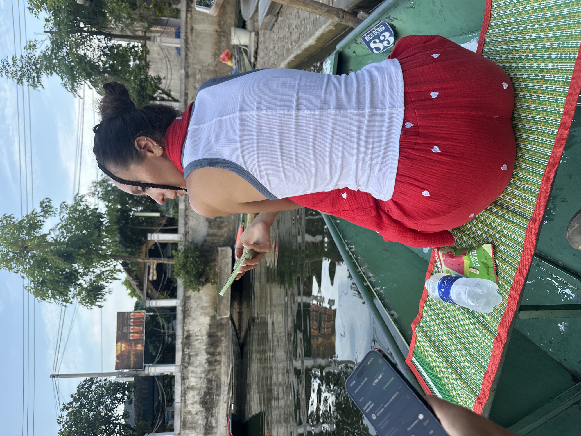 A woman, viewed from behind, sits in a green boat on a waterway in Nam Hoa Lư Ward, Vietnam. She has dark hair styled in a bun with braids and is wearing a white tank top with grey trim and a red skirt adorned with small white hearts. In her right hand, she holds a light green smartphone. In front of her, on a green and yellow woven mat, are a bottle of Aquafina water and a red and white packaged snack with Vietnamese text and an image of a watermelon slice. Another person's hand holds a black smartphone in the lower left corner of the image. The boat is on a body of water with reflections of trees and buildings on the surface. In the background, a concrete wall runs along the bank, topped with trees. A sign with images of beer glasses is visible on the wall. The sky is overcast. A blue and white sticker with the words "BÍCH ĐỒNG" and a stylized "88" is affixed to the side of the boat near the woman's right hip.