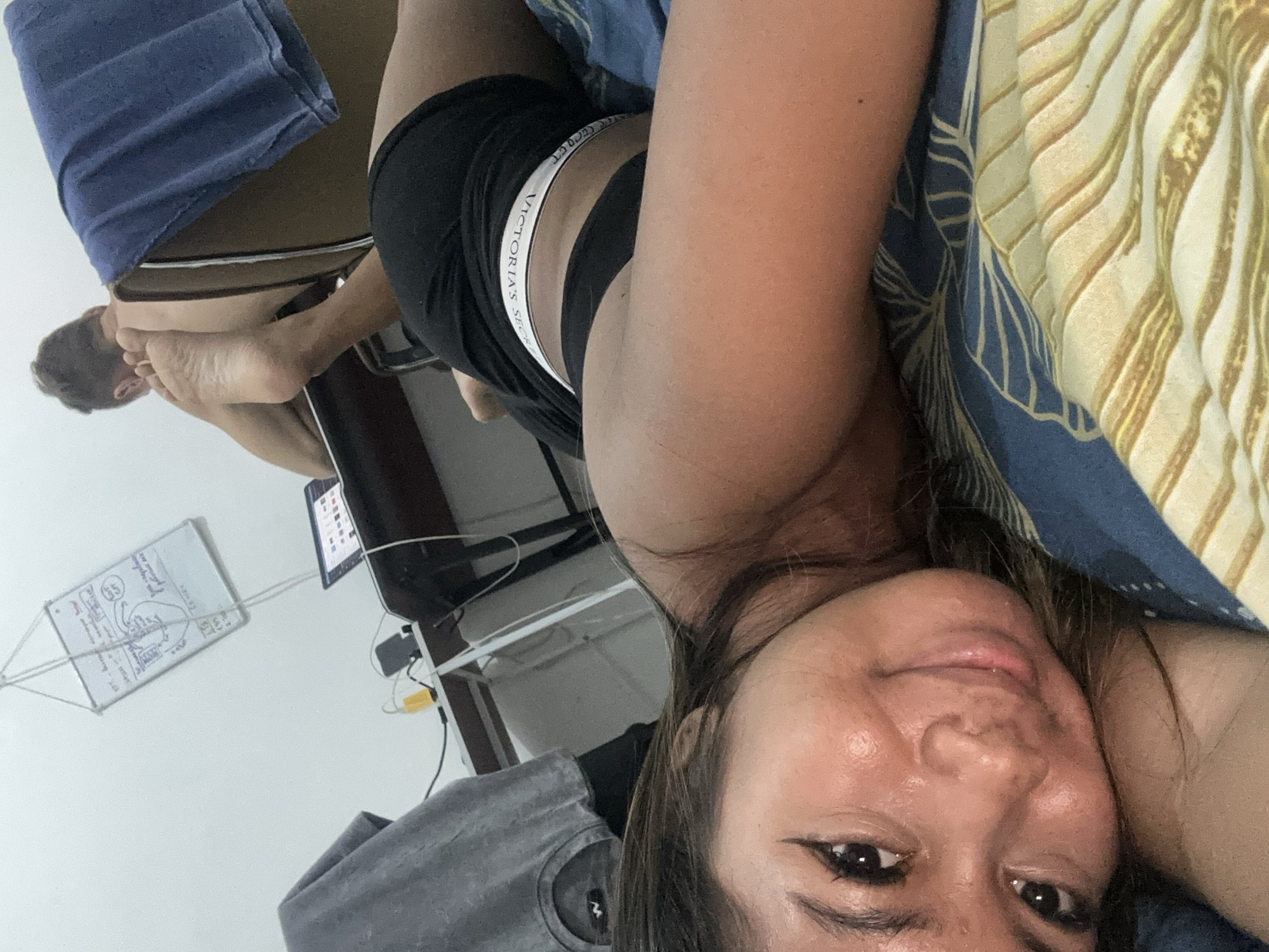 In a room with light-colored walls, a woman is lying on a bed with blue and yellow patterned bedding, facing the camera. She is looking at the camera with a slight smile. Her face is illuminated by the overhead light, highlighting her make-up and skin. Her hair is dark brown and falls around her face. She is wearing black shorts with a white waistband.
Behind her, a man is sitting at a desk with a laptop. He is leaning forward, with his back to the camera. His legs are extended and propped up on the chair. A blue towel is draped over the back of the chair. A whiteboard with writing and diagrams is hanging from the ceiling. Various cables and a yellow object are on the desk. The room appears to be a bedroom or living space, likely in Sukawati, Indonesia, given the context.