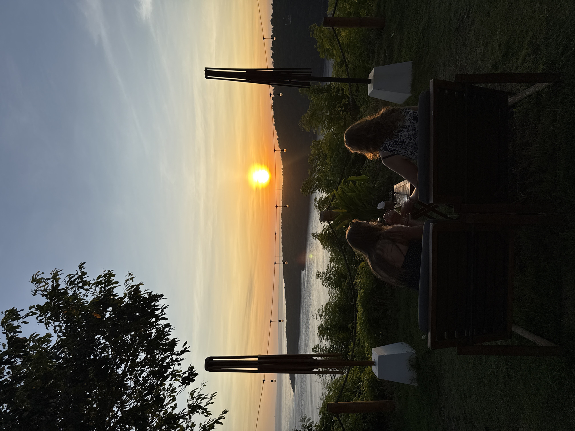 Here's a description of the image:
Two people are seated in chairs at a scenic overlook at sunset. The location is on Nusa Penida, Indonesia, as evidenced by the typical tropical environment and landscape. The sun is setting over the water, casting a golden hue across the sky. String lights hang across the scene, and the water is visible in the distance. The seating area has a relaxed ambiance. The people are engaged in some interaction, most likely enjoying the view. One distinct element is an elongated decorative wind chime or sculptural art piece next to the chairs. The plants in the foreground suggest a garden-like setting.