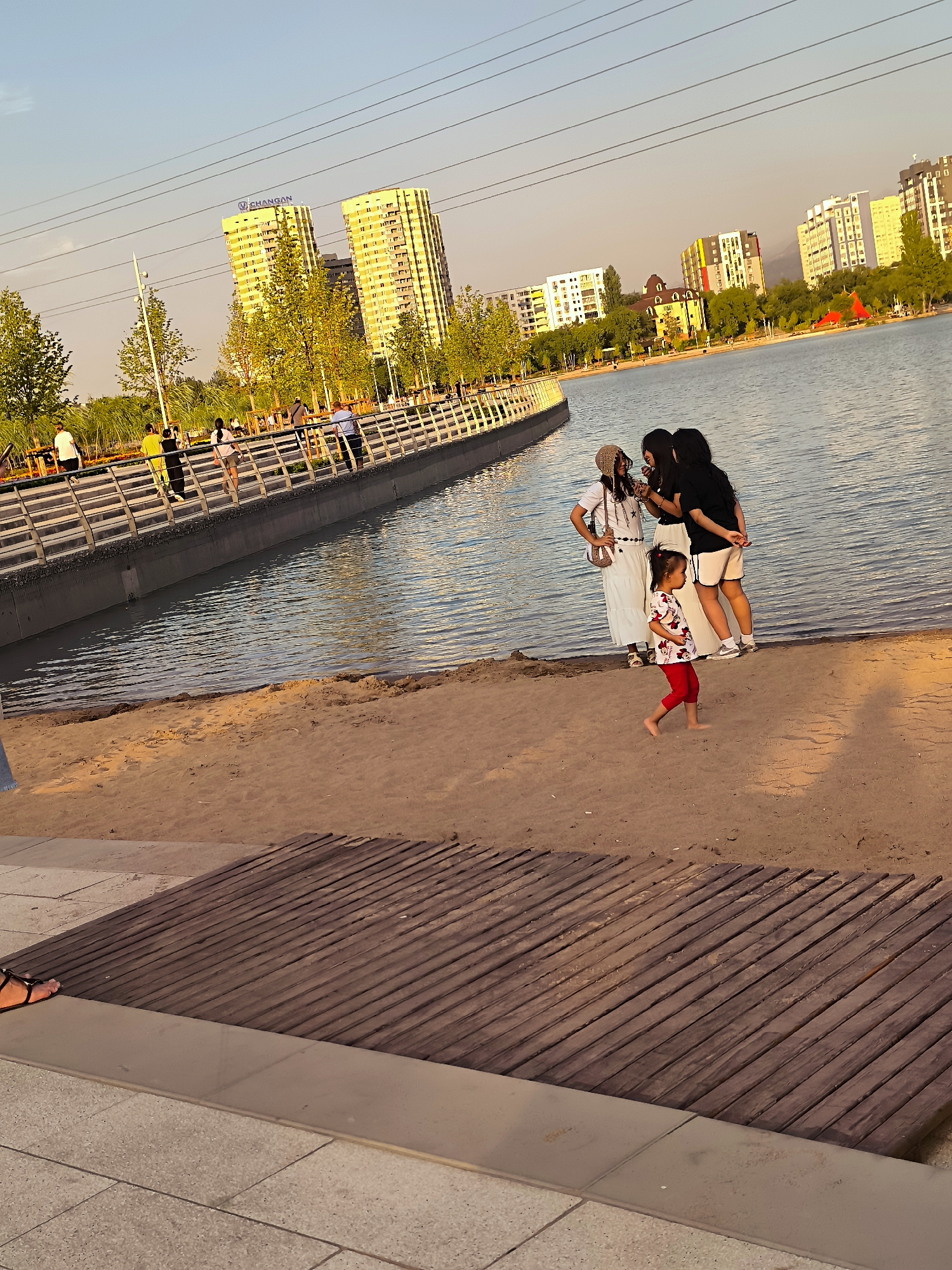 The image captures a serene evening at a waterfront park in Almaty, Kazakhstan, likely during summer. The sun, low on the horizon, casts a warm, golden glow across the scene, illuminating the rippling surface of the artificial lake. In the background, modern high-rise buildings, characteristic of Almaty's urban landscape, rise against a clear, pale blue sky. The buildings are a mix of architectural styles, some featuring vibrant yellow facades, others with more contemporary designs. Trees with lush green foliage line the waterfront, interspersed with walkways and pedestrian bridges.
In the foreground, a sandy beach meets the water's edge. A young child, wearing a red and white patterned outfit, is captured in mid-stride, her bare feet digging slightly into the sand, suggesting playful movement. Further back, near the water, three young women stand together, engaged in conversation. One woman wears a white dress and a straw hat, another a white t-shirt and black shorts, and the third a black t-shirt and white shorts. They appear to be casually socializing, their interaction adding a human element to the picturesque setting.
The park features a well-maintained walkway bordering the lake, with people strolling along it. The walkway is constructed with a modern design, featuring metal railings and a grey concrete base. Further towards the viewer, a section of wooden decking, laid diagonally, forms part of the park's amenities. The overall impression is one of a well-developed urban park offering recreational space and scenic views for its visitors. The presence of high-rise buildings and the lake suggests this is likely a developed residential or commercial area within Almaty.