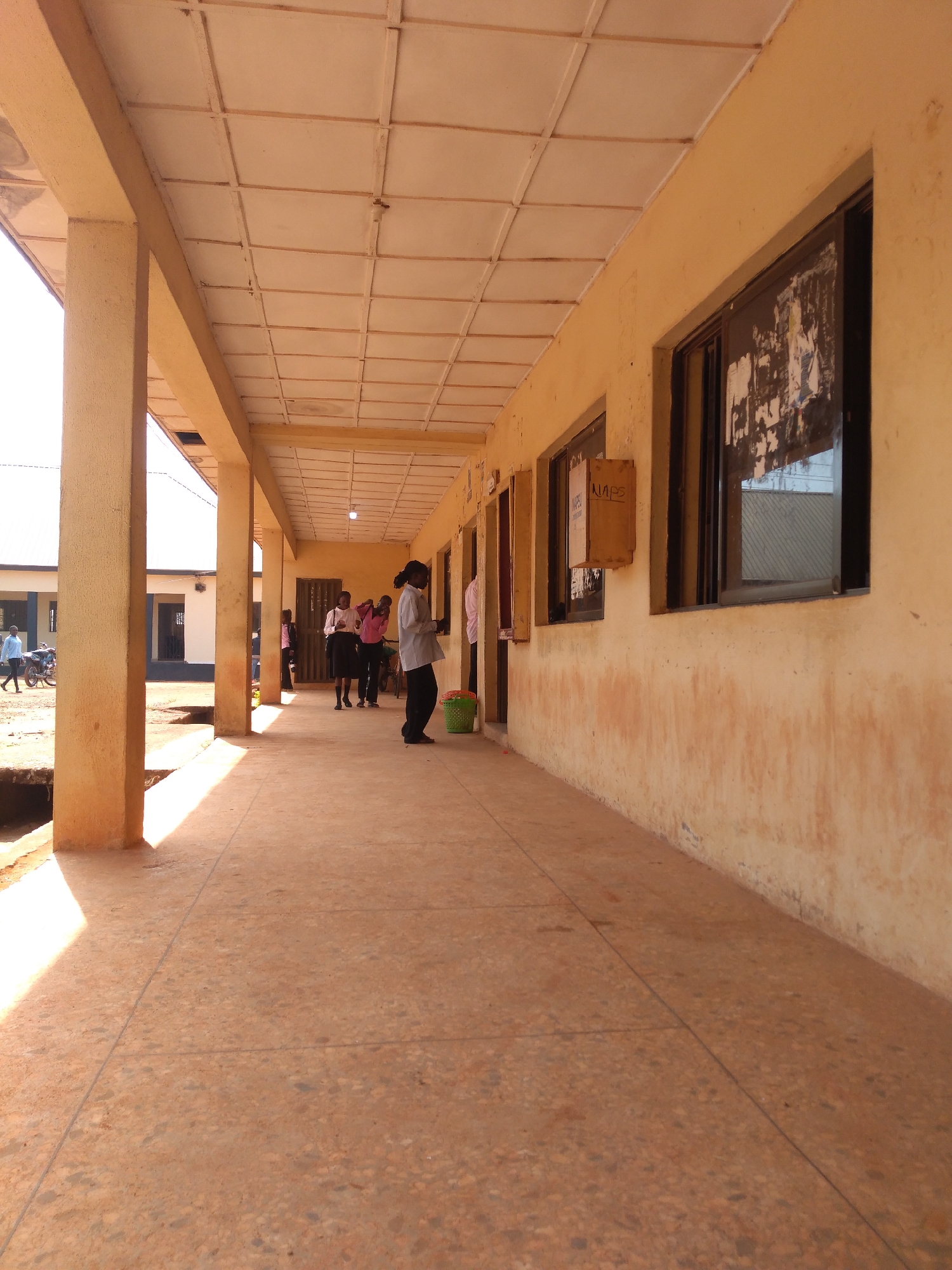 The image depicts an outdoor corridor of a building with a tiled ceiling and yellow-painted walls. Sunlight casts long shadows across the tiled floor. Several individuals, predominantly young women dressed in school uniforms, are visible in the corridor. Some are standing in groups, while others appear to be walking or interacting. One young woman in the foreground is wearing a light-colored long-sleeved shirt and dark pants. A green plastic bucket is placed near her feet. In the background, another building is visible, and a motorcycle is parked to the left. The scene suggests a school or educational institution setting. The location is identified as Jos, Nigeria.