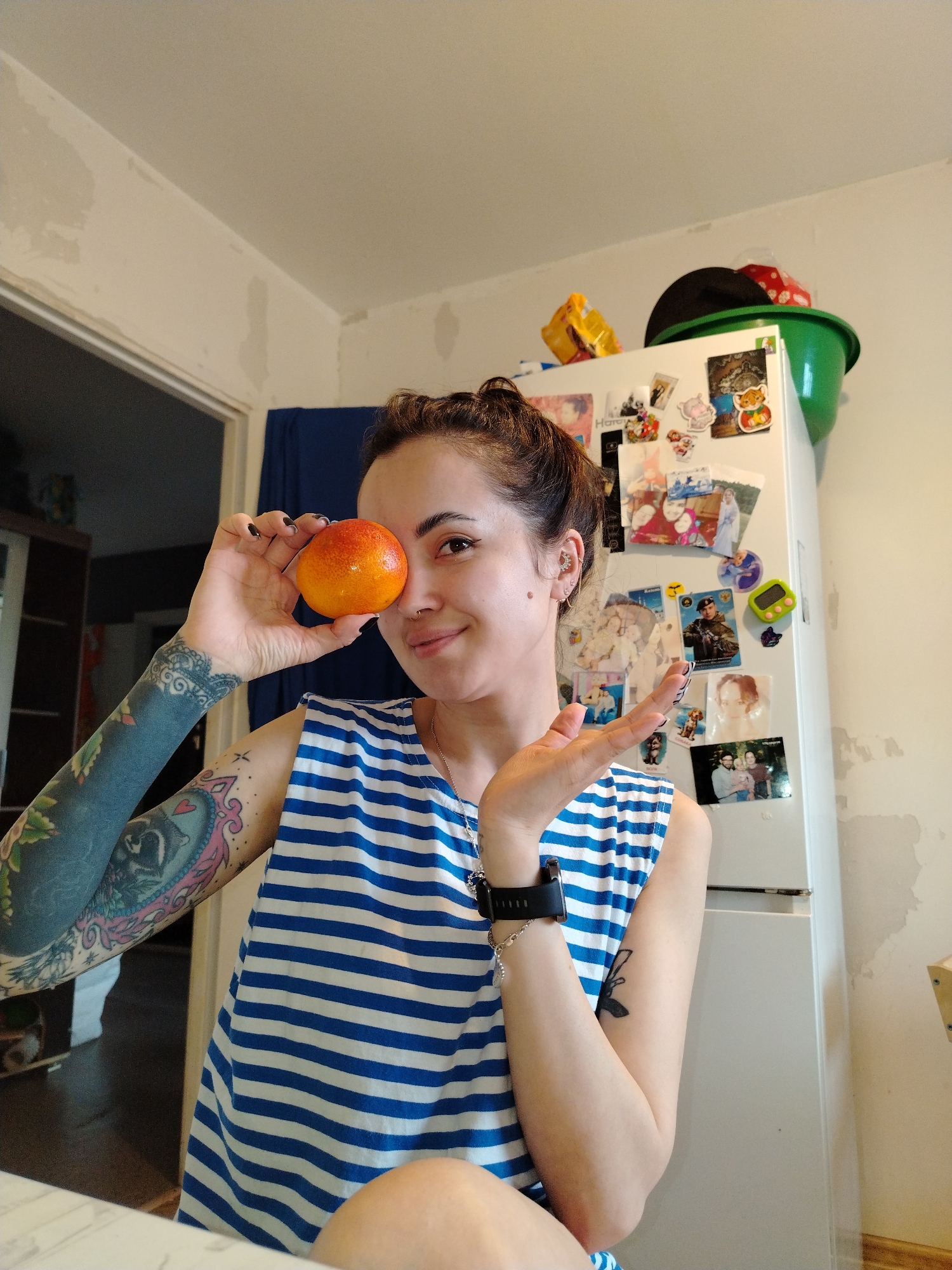 The image shows a young woman playfully posing in what appears to be a home kitchen or living area. She has dark hair pulled back and is wearing a blue and white striped sleeveless top. She holds a round, reddish-orange fruit, possibly a blood orange or persimmon, up to her left eye, looking at the camera with a slight smile. Her left arm is heavily tattooed with a dark sleeve and a colorful framed illustration featuring an animal resembling a raccoon, while her right forearm has a smaller black tattoo. She wears a nose ring, multiple ear piercings, a necklace with a dark pendant, and a black smartwatch on her right wrist.
Behind her, a white refrigerator is covered with various magnets and personal photos. On top of the refrigerator, a green plastic bowl, a black hat, and other items are stored. The light-colored walls behind the woman and to the right of the refrigerator show patches of what looks like exposed plaster or peeling paint. A blue curtain or fabric is visible in the background, to the left of the refrigerator, and a doorway is also visible on the left side of the frame. In the foreground, a light-colored countertop is present. The visible text on one of the refrigerator magnets appears to be "HOTO." The overall atmosphere is casual and cheerful, with the woman's expression conveying friendliness.