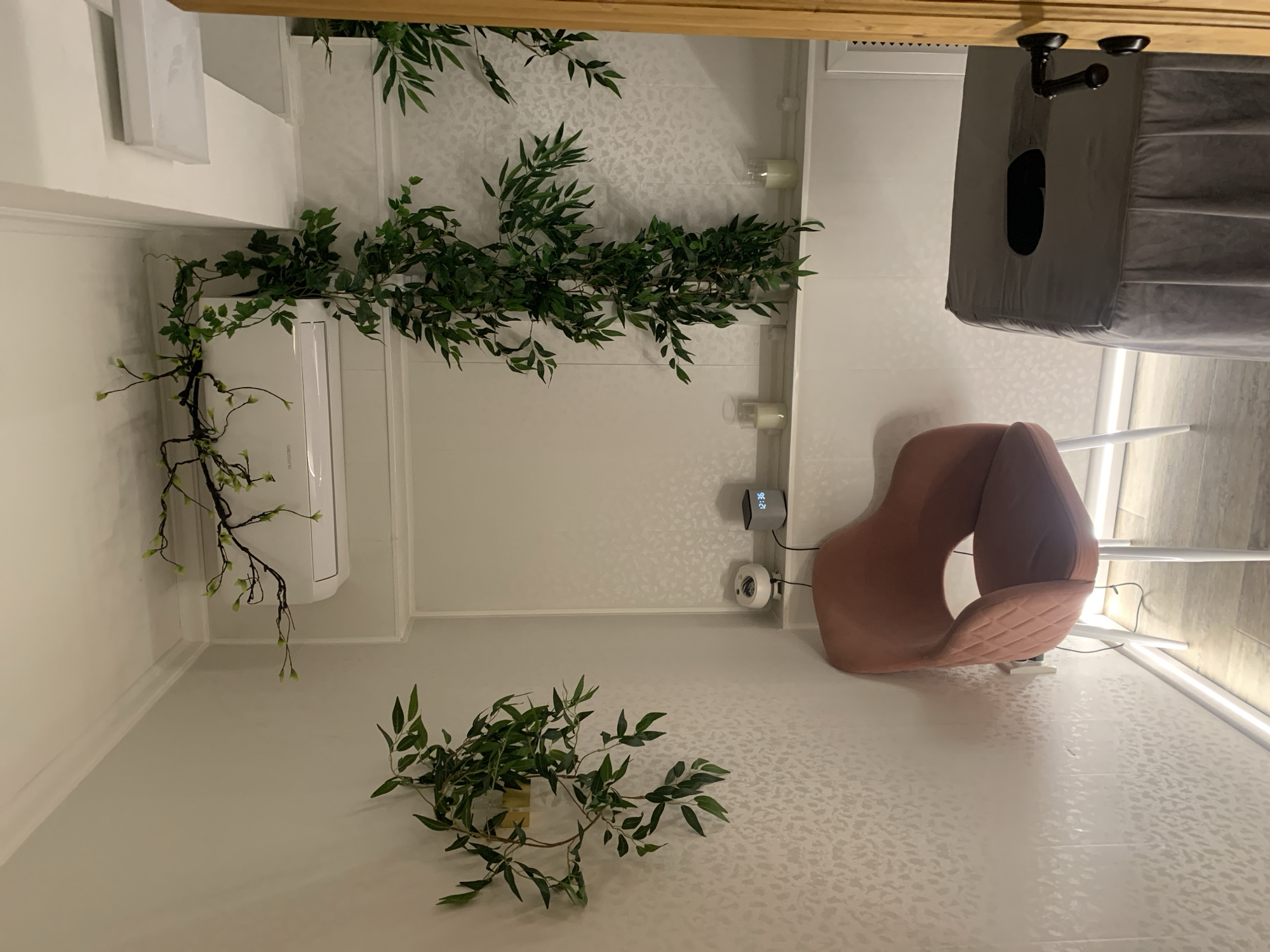 The image depicts an indoor spa or treatment room. The walls are painted a neutral, light color with a textured pattern. A white air conditioning unit is mounted high on the wall, draped with artificial green vines. Two large potted plants with green foliage are positioned on the left side of the room, one hanging and one on a shelf.
In the center of the room, a plush, pink velvet armchair with white legs is placed. To the right of the armchair, a white shelf holds a small white humidifier and a silver digital clock displaying "12:36". Further to the right, on the same shelf, are two glass candle holders with lit white candles. A large, gray massage table covered with a fitted sheet and a dark rectangular opening for the face is situated in the foreground on the right side of the image. A wooden door with a black handle and lock is partially visible on the far right.
The lighting appears to be soft and ambient, possibly from recessed ceiling lights or wall sconces not fully visible. The floor is tiled with a wood-grain pattern. The overall atmosphere suggests a calm and relaxing environment. The location is identified as Almaty, Kazakhstan.