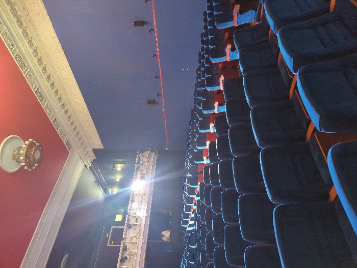 The image depicts the interior of the main auditorium at the Khudozhestvenny Cinema in Moscow, Russia. The perspective is from the front section of the ground floor seating, looking towards the stage/screen area and up towards a balcony on the left.
The foreground and midground are dominated by numerous empty rows of dark blue velvet seats featuring wooden armrests. Some seats in the distance, particularly when folded, reveal red undersides. The side walls of the auditorium are painted a uniform deep blue, contrasting with a deep red ceiling. A white, ornate crown molding separates the walls and ceiling, extending along the perimeter. A large, ornate, gold-toned chandelier with multiple light fixtures hangs centrally from the red ceiling.
On the left side, a white, ornate balcony railing is visible, with a strong, bright light source emanating from that level, causing a significant lens flare. Below this light source and the balcony, a single individual wearing a light-colored shirt is partially visible, appearing to be standing or seated at a console. On the right blue wall, two small, dark speakers are mounted at mid-height. Below them, a horizontal red line or strip runs across the wall, adorned with several small, dark, hook-like fixtures. The overall lighting is dim, with the bright light source from the balcony being the most prominent illumination.