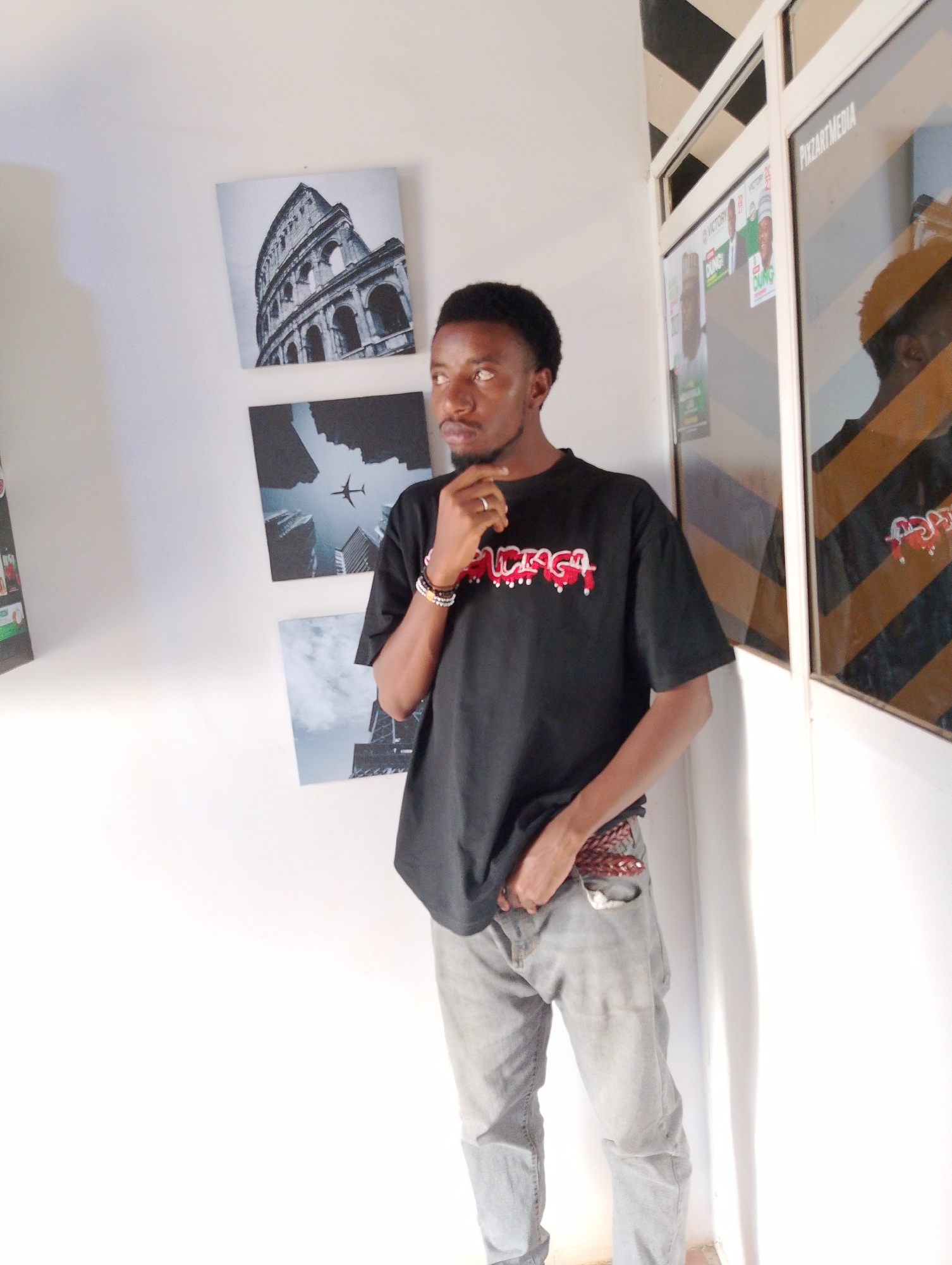 A young Black man stands with his right hand on his chin, looking to the left of the frame. He has short, dark, curly hair and is wearing a black t-shirt with red and white lettering that reads "LURAGA," and light-wash jeans with a patterned belt. He is standing in a well-lit, indoor space with a white wall to his left. On the wall, there are three square pictures. The top picture shows the Colosseum in Rome. The middle picture shows an airplane flying over skyscrapers. The bottom picture is of skyscrapers. To the man's right is a glass-paneled doorway with black and yellow striped markings. Through the glass, political posters with images of men and the word "VICTORY" and "DUGUNG" are visible. A reflection of the man and a portion of the interior is visible in the glass. The environment appears to be a commercial or office space, possibly in Jos, Nigeria, given the presence of political posters common in Nigerian elections.