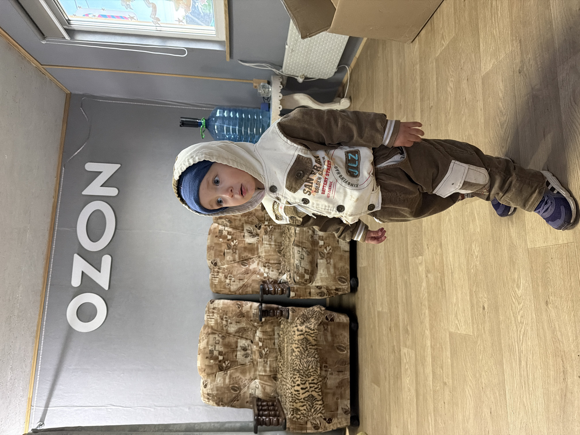 A toddler, wearing a blue beanie, a hooded brown and white jacket with patches that say "SAN FRAN GOLDEN GATE BATTERY STREET 18TH AVENUE JLZ," and brown pants, stands on a light-colored wooden floor. The toddler is looking towards the camera with a curious expression. Behind the toddler are two brown armchairs with a patterned upholstery. To the left of the toddler, on a gray wall, are large white letters that spell out "OZON." Further in the background, on a shelf, is a large blue water jug. The scene appears to be indoors, likely in a room with a window at the top left corner, suggesting it might be daytime. There are no overt indications of the weather. The location is identified as Красный Маяк, Russia.