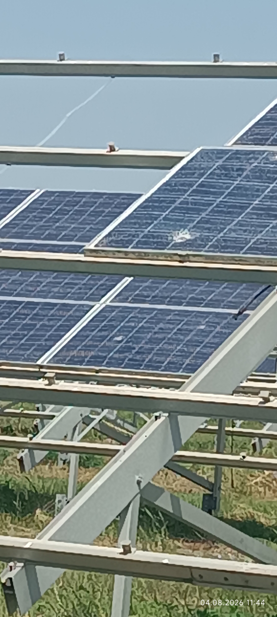 The image shows a close-up view of a solar panel array under a clear sky. The solar panels are arranged in multiple rows, angled upwards and supported by a metallic structure. The structure consists of horizontal beams and vertical supports, all made of a light-colored metal, likely aluminum or galvanized steel. The ground beneath the panels is covered with dry, green and brown vegetation, suggesting an outdoor, possibly rural, location. There are some small, dark objects visible on the top beams, possibly fasteners or debris. The overall impression is one of infrastructure for renewable energy generation. A timestamp "04.08.2026 11:44" is present in the bottom right corner of the image. The location is identified as Starokozache, Ukraine.