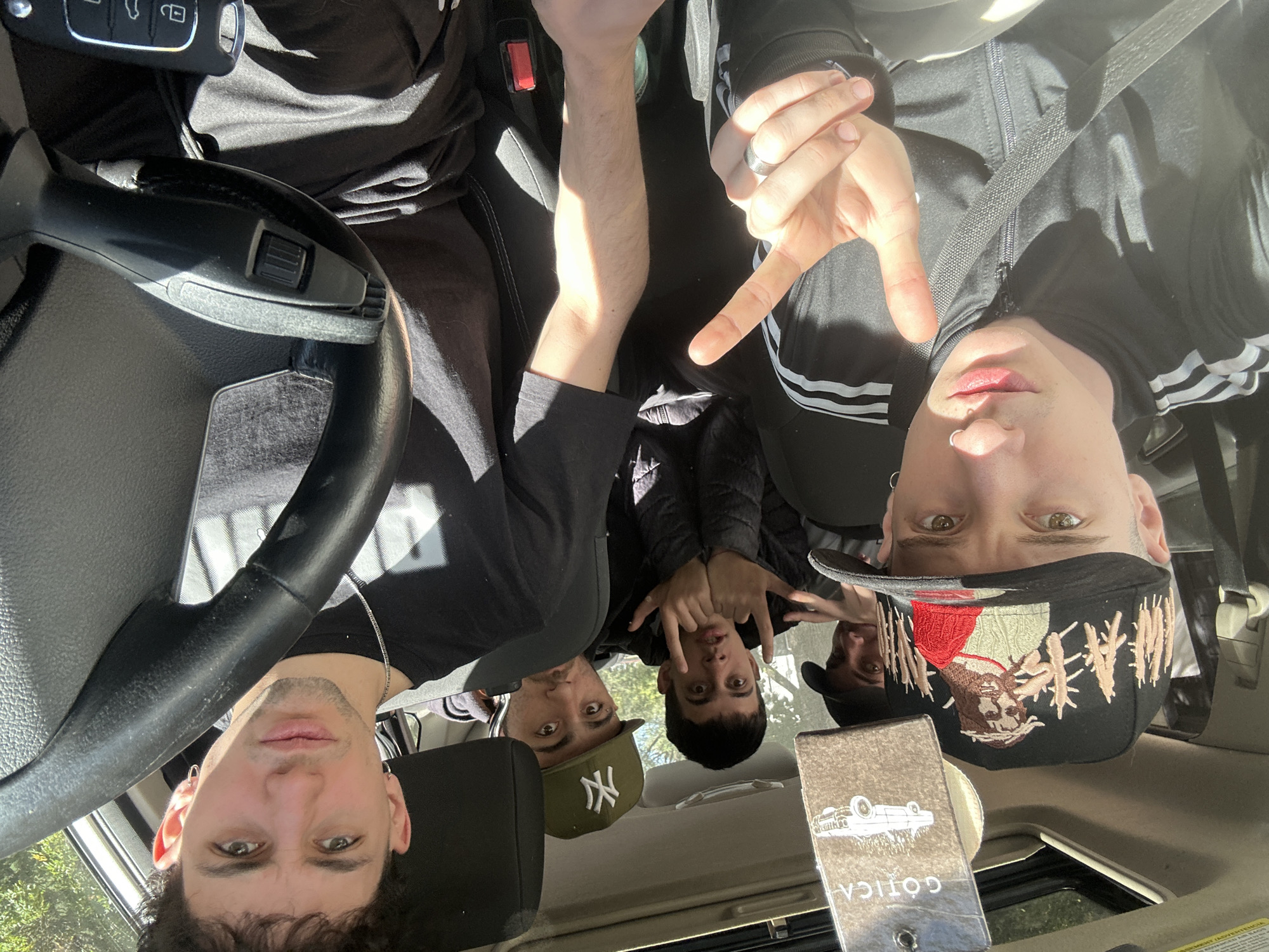 Four young men are captured in a casual selfie taken inside a car during the daytime. The strong sunlight casts prominent shadows, indicating a clear and sunny day. The image appears to be taken from a slightly elevated perspective, looking down into the car.
In the foreground, on the left, a man with dark, curly hair and light eyes looks directly at the camera. He is situated near the black steering wheel of the car. In the front passenger seat, another man wearing a black Adidas jacket with white stripes makes a hand gesture with his right hand, potentially a peace sign or "V" gesture, and has a nose piercing. His left arm is visible, illuminated by sunlight.
In the back seat, two more young men are visible. The one on the left wears a green baseball cap with a white "NY" logo. The man on the right, also looking at the camera, wears a black baseball cap featuring a distinctive graphic of a figure and some text, which is partially obscured. He is also making a hand gesture.
Various objects are visible within the car: a set of car keys with an Audi logo, seatbelts, and a metal air freshener or pendant hanging from the rearview mirror area. This pendant has "GOTICA" written vertically on it and features an illustration of an overturned car. The car's interior appears clean, with black upholstery. The activity seems to be a group of friends taking a relaxed photo together, possibly while traveling or parked. The overall mood is informal and friendly. The scene is set in Buenos Aires, Argentina.