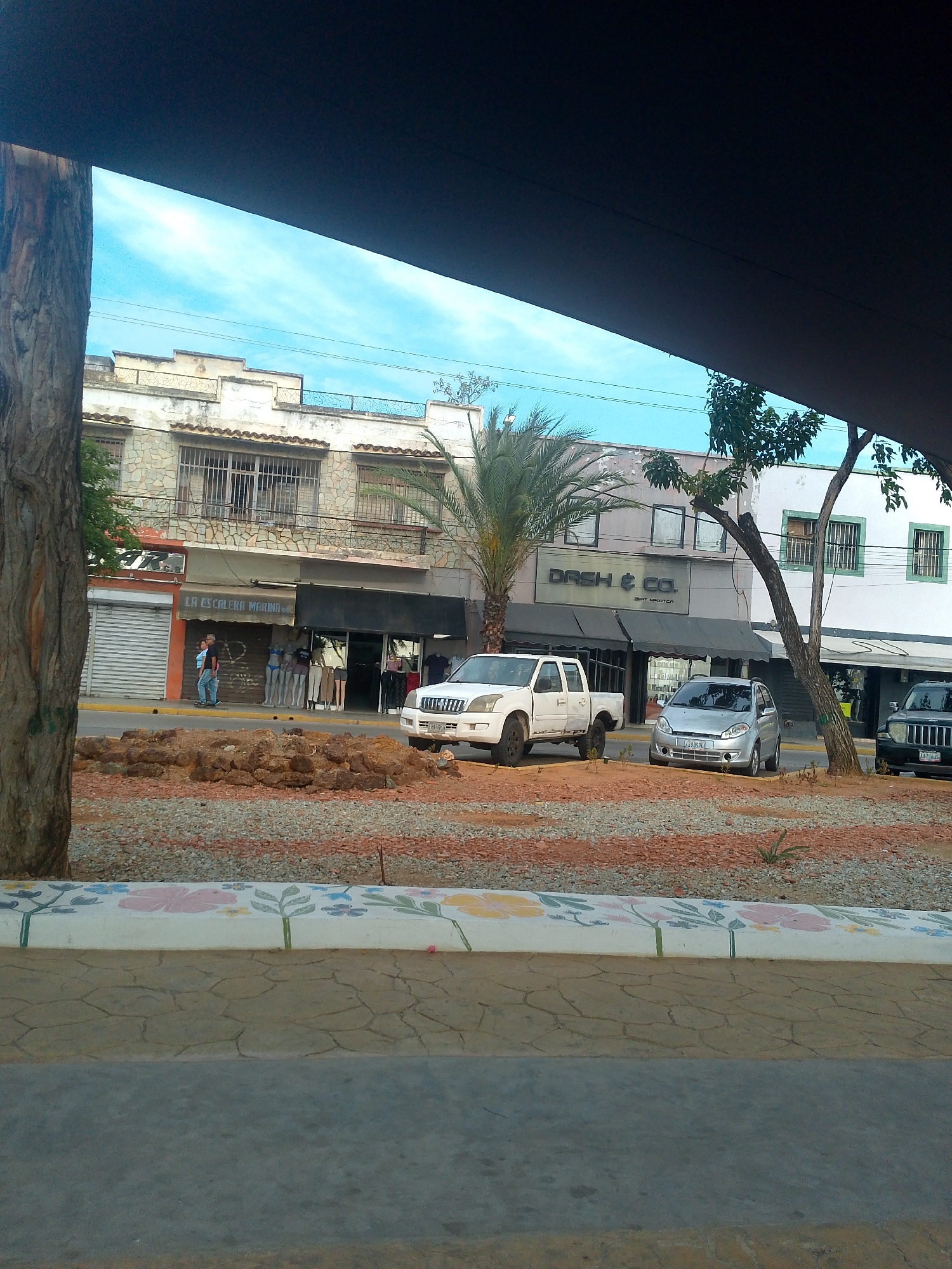 The image depicts a street scene in Juan Griego, Venezuela, focusing on a section of the street lined with two-story buildings. Several vehicles are parked along the street, including a white pickup truck, a silver compact car, and a dark Jeep. The facades of the buildings feature businesses like "LA ESCALERA MARINA" and "DASH & CO." The street is separated from the sidewalk by a median filled with gravel and some larger rocks. A section of the sidewalk features a decorated low wall with painted floral designs. The sky is visible in the upper portion of the image, partially obscured by an overhanging structure, and there are also trees present in the scene. A few people are visible in front of the "LA ESCALERA MARINA" store.