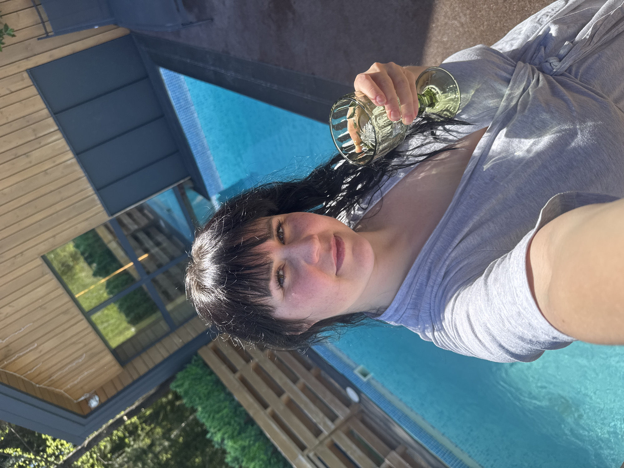 A woman with dark hair and bangs is holding a glass in front of a swimming pool. The background features a wooden structure with windows reflecting greenery. The location appears to be outdoors, possibly in a backyard setting. The image is taken from a slightly upward angle, suggesting a selfie. There is no information available to confirm or deny that the location is in Izhevsk, Russia.