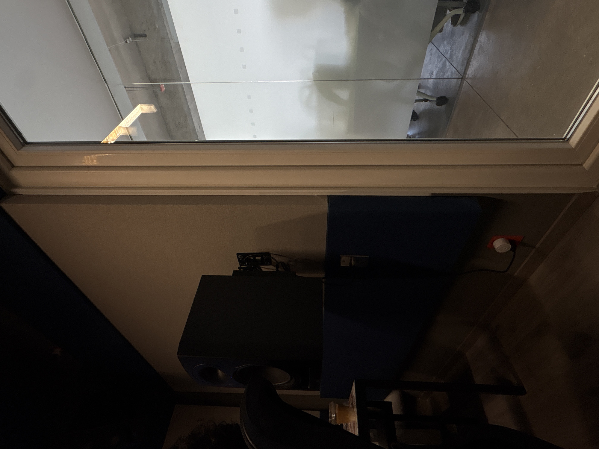 A low-angle indoor shot shows a window view looking out onto what appears to be the interior of a building. The window frame is light-colored, and the glass reveals reflections of the building interior, including indistinct figures of people and furniture such as office chairs. Underneath the window, there's a dark-colored speaker or electronic device placed on a surface. A chair is partially visible in front of the speaker. A red plug is visible on the right. There is not enough information in the image to identify the location in Mexico City, Mexico.