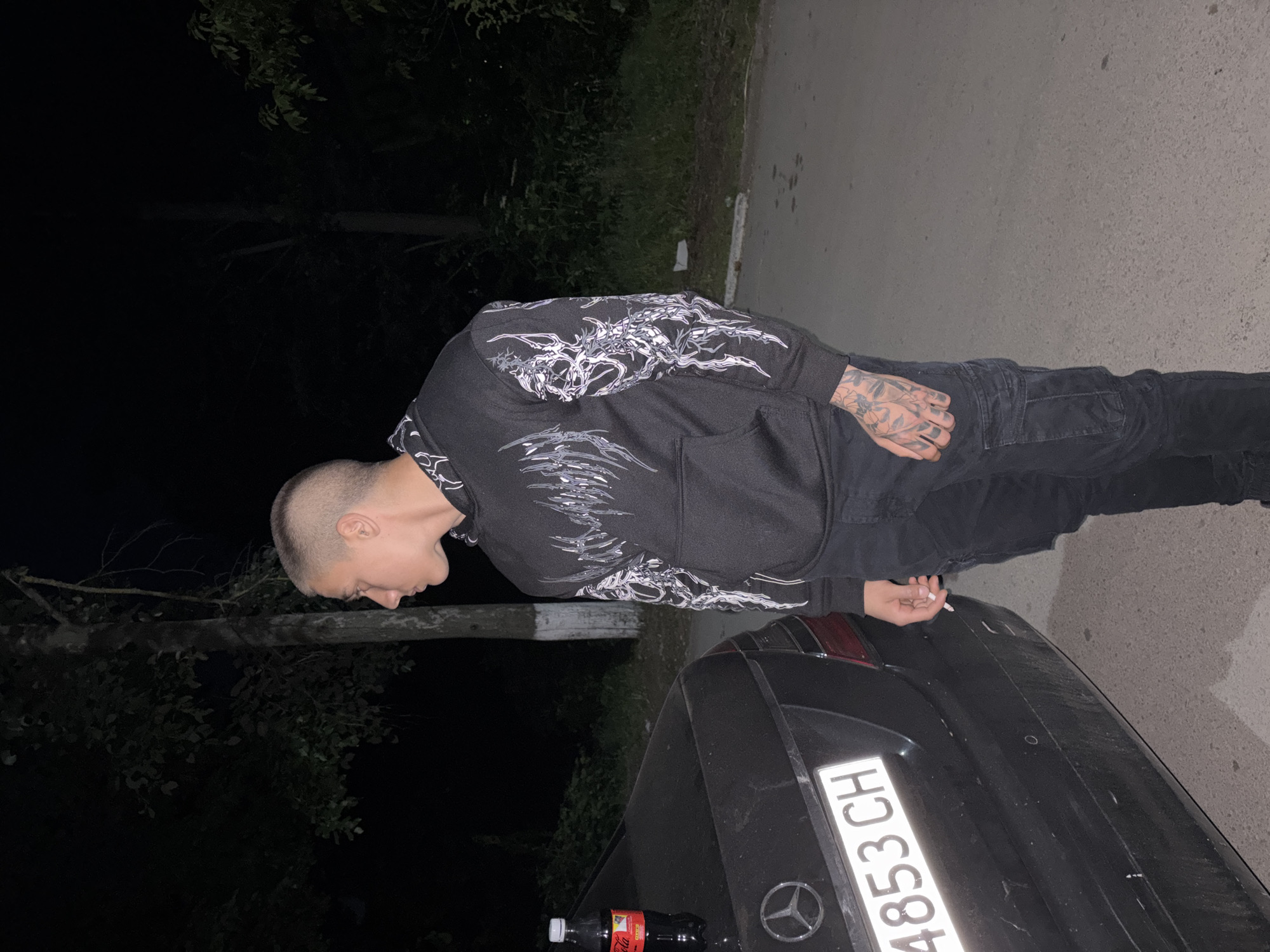 A person with a shaved head, wearing a dark hoodie with a white design and dark pants, stands next to a dark-colored car. The car's license plate reads "4853 CH". A bottle of Coca-Cola sits on the trunk of the car. The setting is a poorly lit area near some trees, possibly in a street. No information about the location can be determined beyond the car's license plate potentially indicating a location in Switzerland. No additional information regarding the city of Mihailovo, Bulgaria, can be determined from the image. The person is looking down at the car. The person is holding a cigarette in their hand.