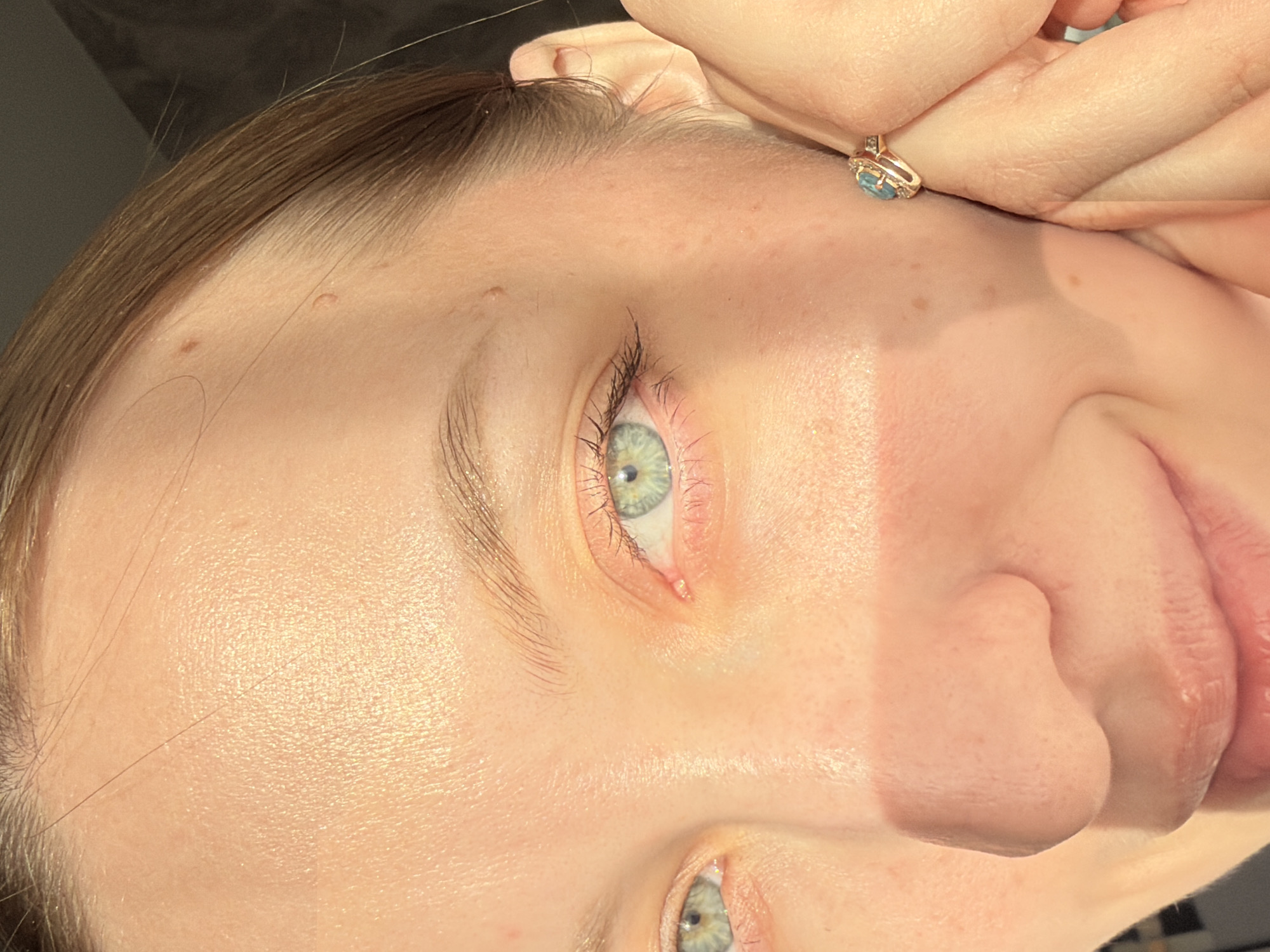 A close-up image shows a person's face. The person has fair skin, light brown hair, and light green eyes. A gold ring with a blue stone is on their finger. The background is dark and out of focus. No evidence suggests this image was taken in Reshetnikovo, Russia.