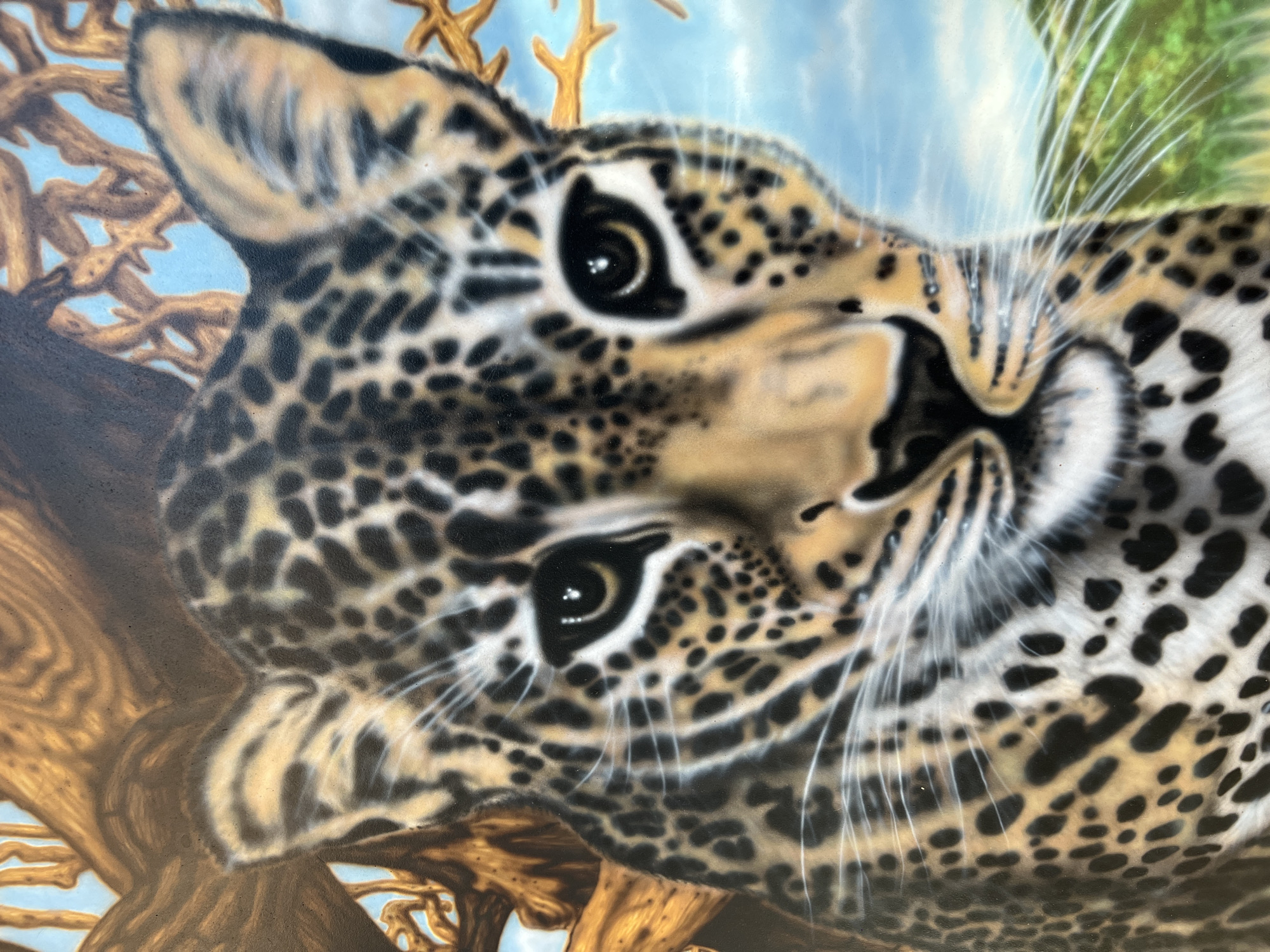 The image is a close-up painting of a leopard's face, rendered in an airbrushed style. The leopard has distinctive black spots on a tawny coat and bright, alert eyes. Behind the leopard, there is a blurred background depicting tree branches against a blue sky and a hint of green foliage. The painting appears to be on a flat surface, likely a wall or a canvas. The image does not depict any specific location within the city of Bender, Moldova, or any people.