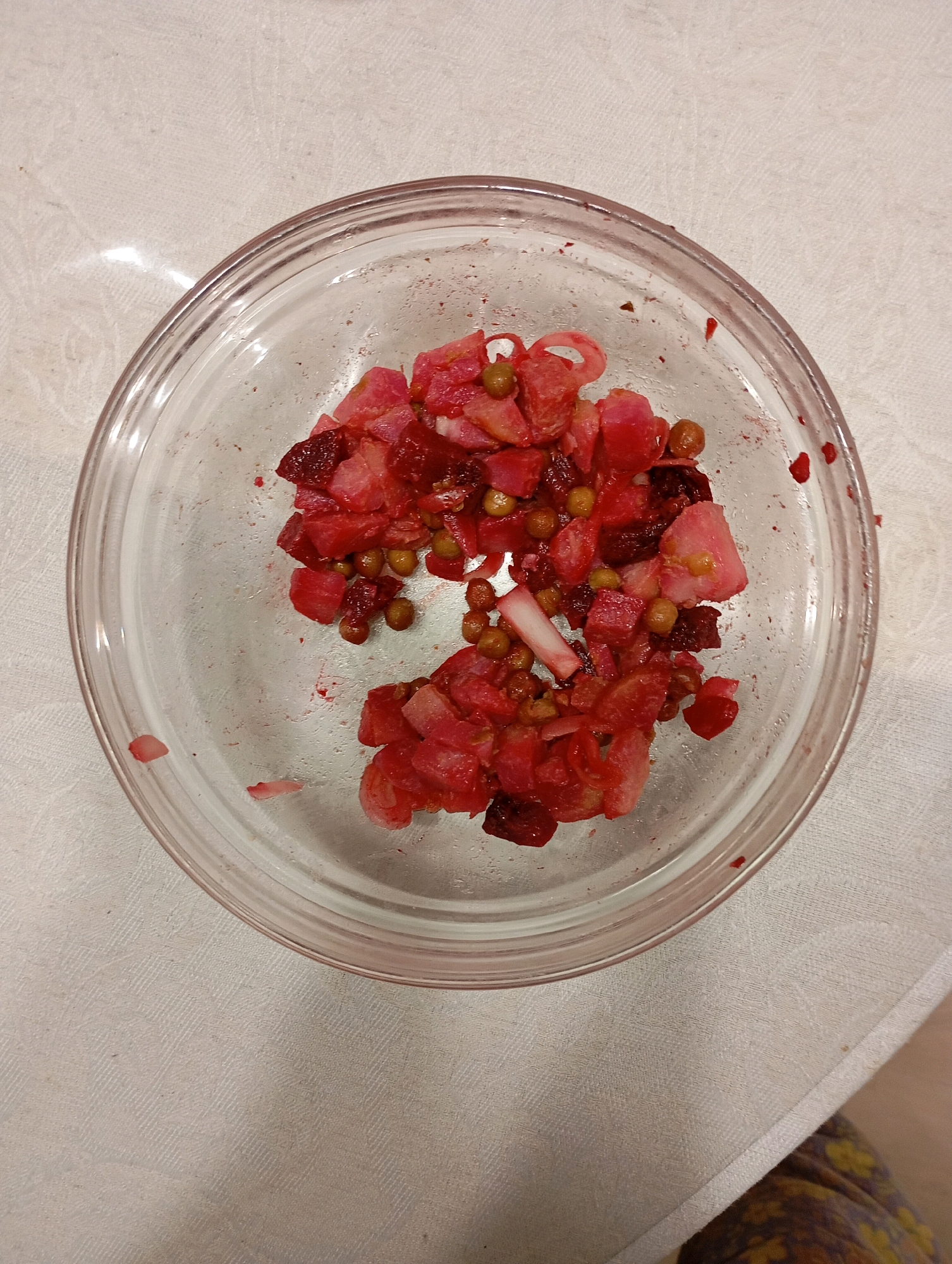 A clear glass bowl containing a portion of Vinaigrette salad is centered on a textured white tablecloth. The salad, a traditional Russian dish, consists of diced cooked beets, potatoes, carrots, peas, and possibly pickles or onions, all mixed and stained red from the beets. Some of the salad has spilled over the edge of the bowl onto the tablecloth. The lighting suggests an indoor setting, likely a kitchen or dining area. The image does not provide any specific geographical markers to indicate its location in Yakutsk, Russia.