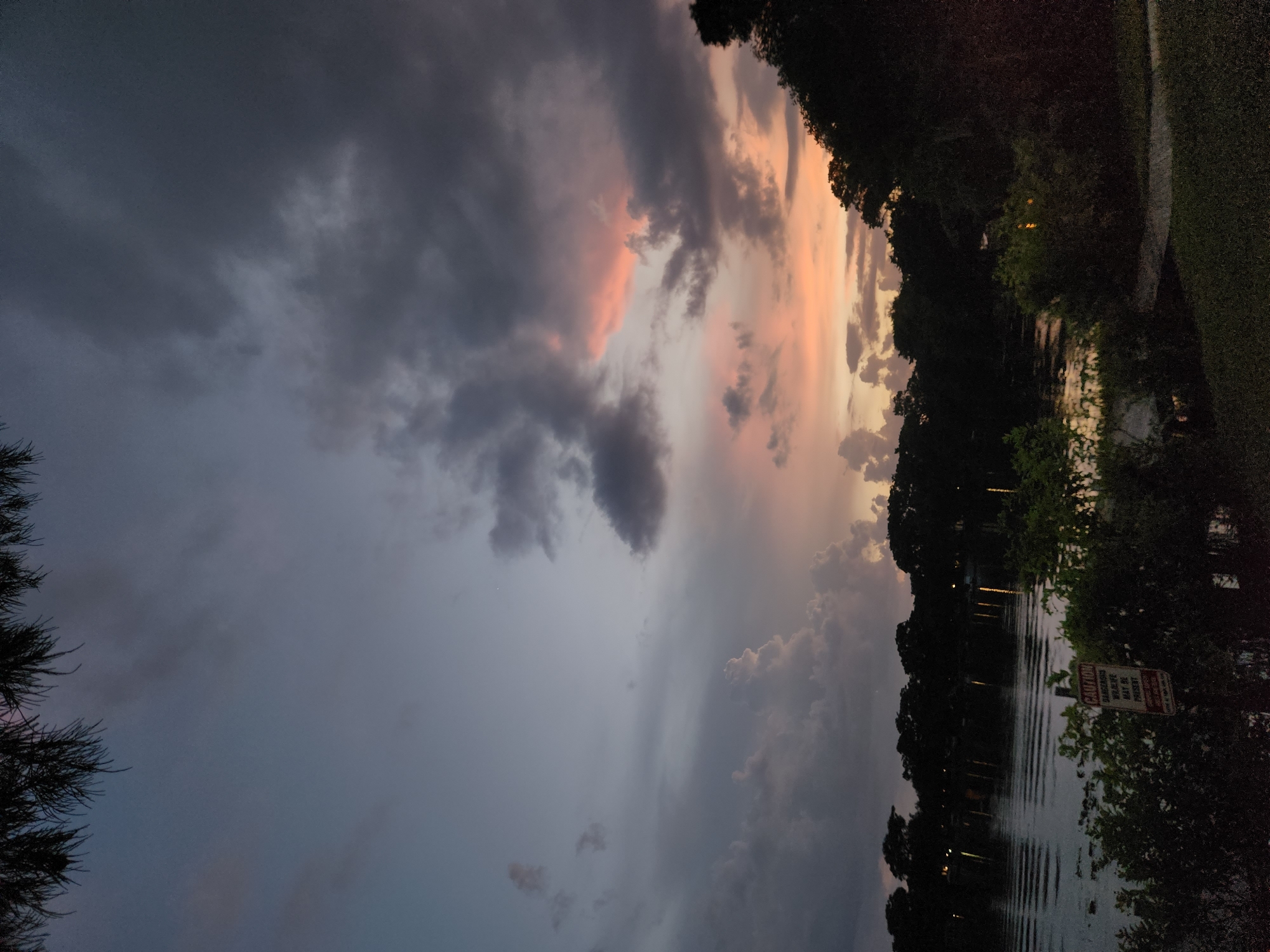 The image depicts a twilight scene over a body of water, likely a lake or canal, bordered by dense foliage and silhouetted trees. The sky is dominated by dramatic clouds, displaying hues of gray, purple, and soft pinks and oranges from the setting sun. In the foreground, there is a sign with red lettering on a white background, partially obscured by vegetation. The text on the sign appears to warn of potential dangers, with the word "CAUTION" clearly visible, followed by smaller text that might read "DANGEROUS WILDLIFE MAY BE PRESENT." The water's surface reflects the sky and the distant lights from what appears to be structures or pathways along the opposite bank. The overall atmosphere is serene and slightly somber due to the fading light. The location is specified as being in Winter Park, United States of America, suggesting a natural or park-like setting within a city.