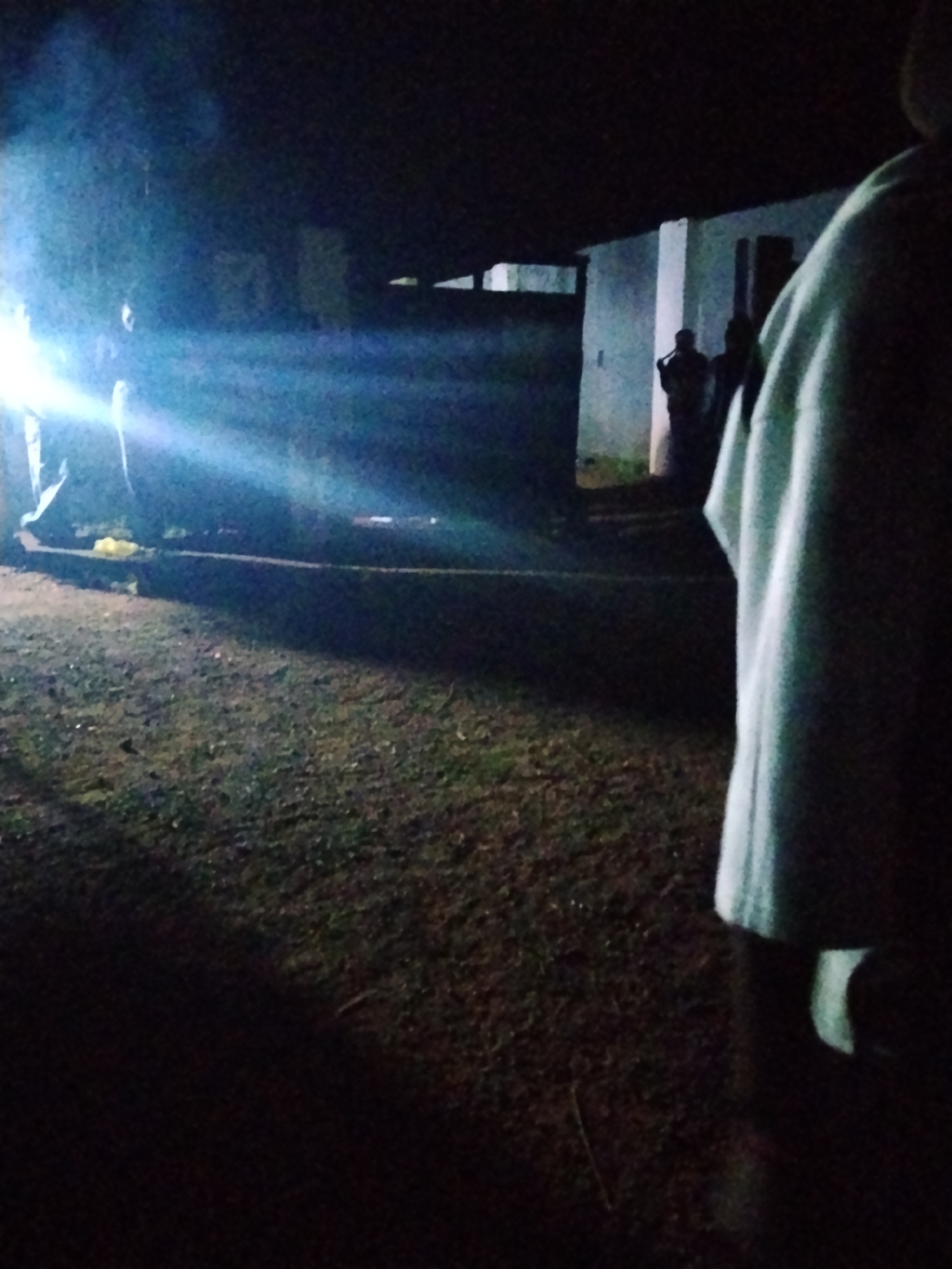 In the city of Durumi, Nigeria, a dimly lit outdoor scene unfolds. The foreground is dominated by the indistinct shape of a person in a white garment, partially obscuring the view. Beyond, a patch of dark, uneven ground suggests a yard or unpaved area. A strong beam of light emanates from the left side of the frame, cutting across the scene and illuminating a low wall or barrier. Behind this barrier, two or three figures are visible in the shadows, standing near a light-colored building with dark openings that could be windows or doorways. The overall atmosphere is dark and somewhat mysterious, with the artificial light creating dramatic shadows and highlights.