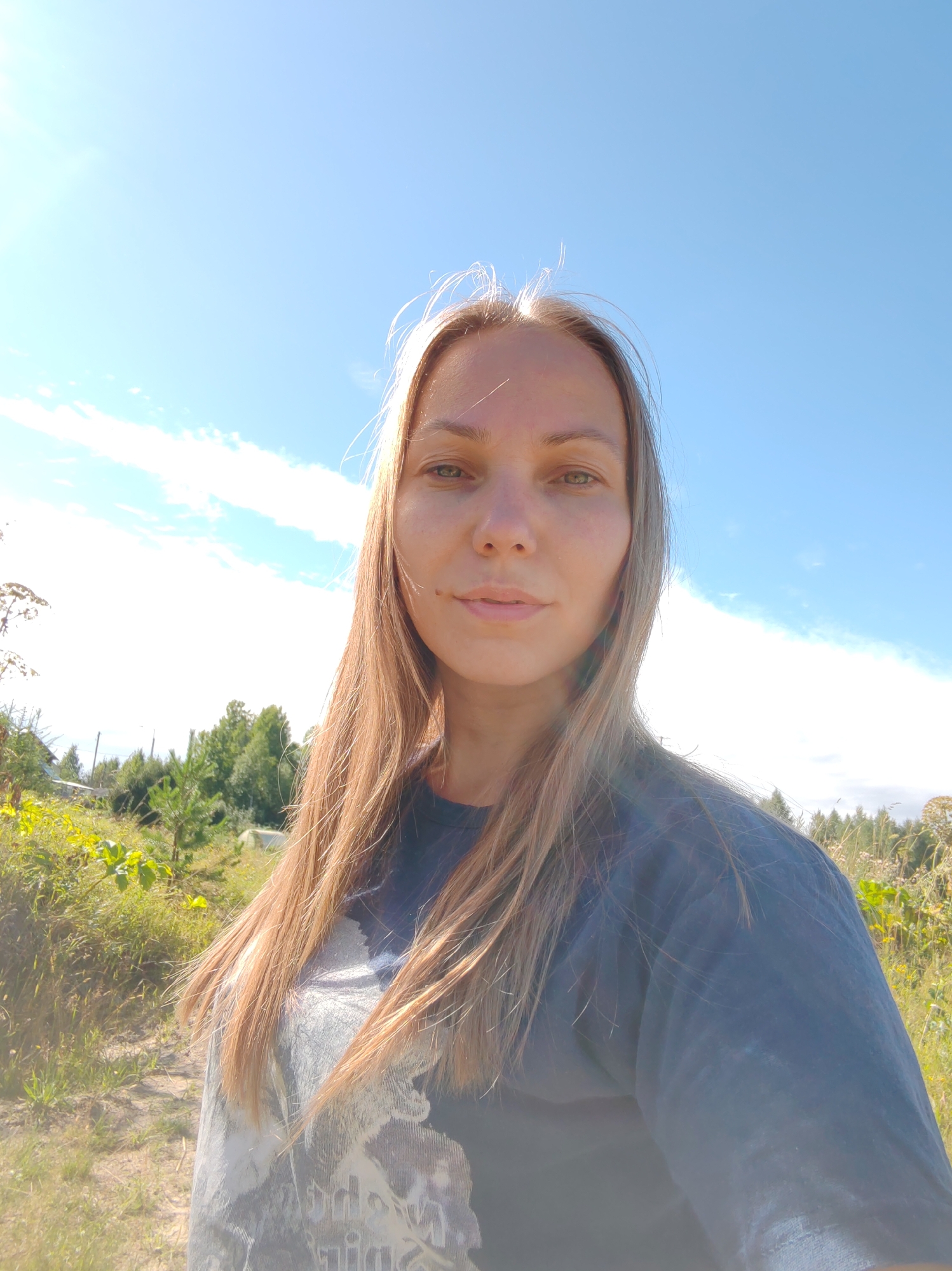 A medium shot selfie features a woman with blonde hair and green eyes, looking directly at the camera. She is outdoors, with a bright blue sky and scattered clouds visible in the background. To her left, there is a lush green landscape with trees and yellow wildflowers. To her right, the sky continues, with hints of trees in the distance. The woman is wearing a dark t-shirt with a graphic print. The lighting suggests a sunny day. The location is described as Chukhlem, Russia, indicating a rural or natural setting.