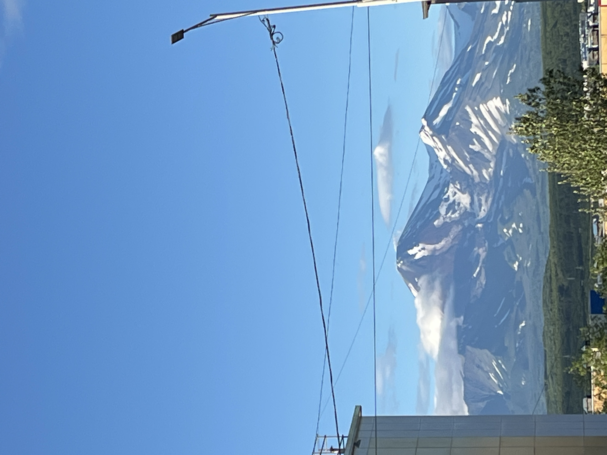 The image displays a view of the Avachinsky and Koryaksky volcanoes in Petropavlovsk-Kamchatsky, Russia, seen from a distance. The foreground features a modern building with a light-colored facade on the left, and a lamppost with a dark rectangular light fixture on the right. Power lines stretch horizontally across the frame, dividing the sky from the mountain landscape. The mountains are partially covered in snow, with the Avachinsky volcano appearing more prominent and closer, while Koryaksky is visible behind it. Patches of clouds are present around the mountain peaks. Below the mountains, a dense green forest covers the landscape, with hints of urban structures visible at the bottom of the image. The sky is a clear, bright blue.