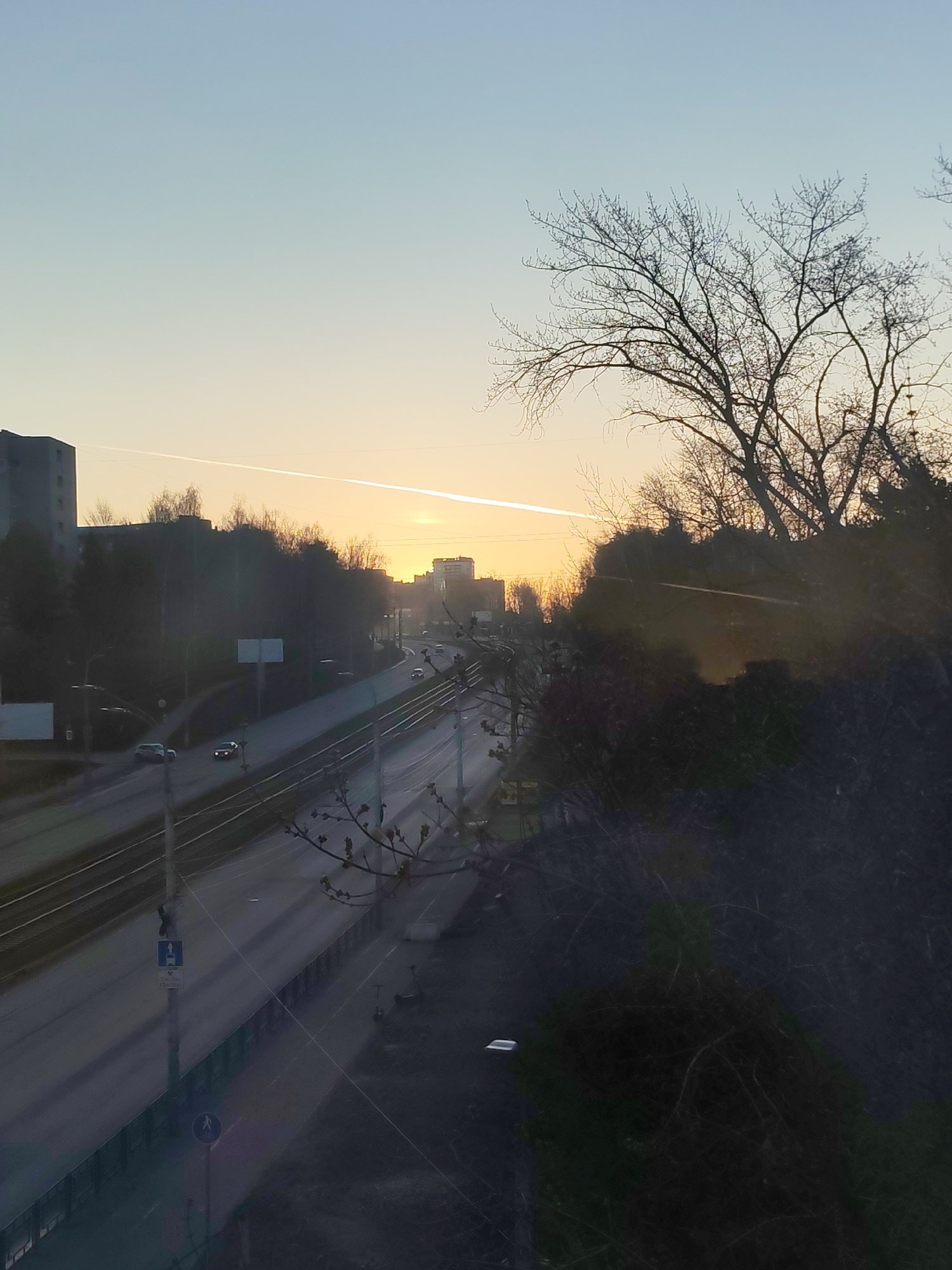 The image shows a cityscape in Izhevsk, Russia, at dawn or dusk. A wide road with multiple lanes and overhead tram lines stretches into the distance. Several cars are visible on the road. Buildings line the horizon, silhouetted against a pale sky. A bright streak, likely a contrail, cuts across the sky. A bare tree stands to the right of the frame, its branches contrasting with the sky. Part of an embankment obscures some of the scenery. The overall tone is peaceful.