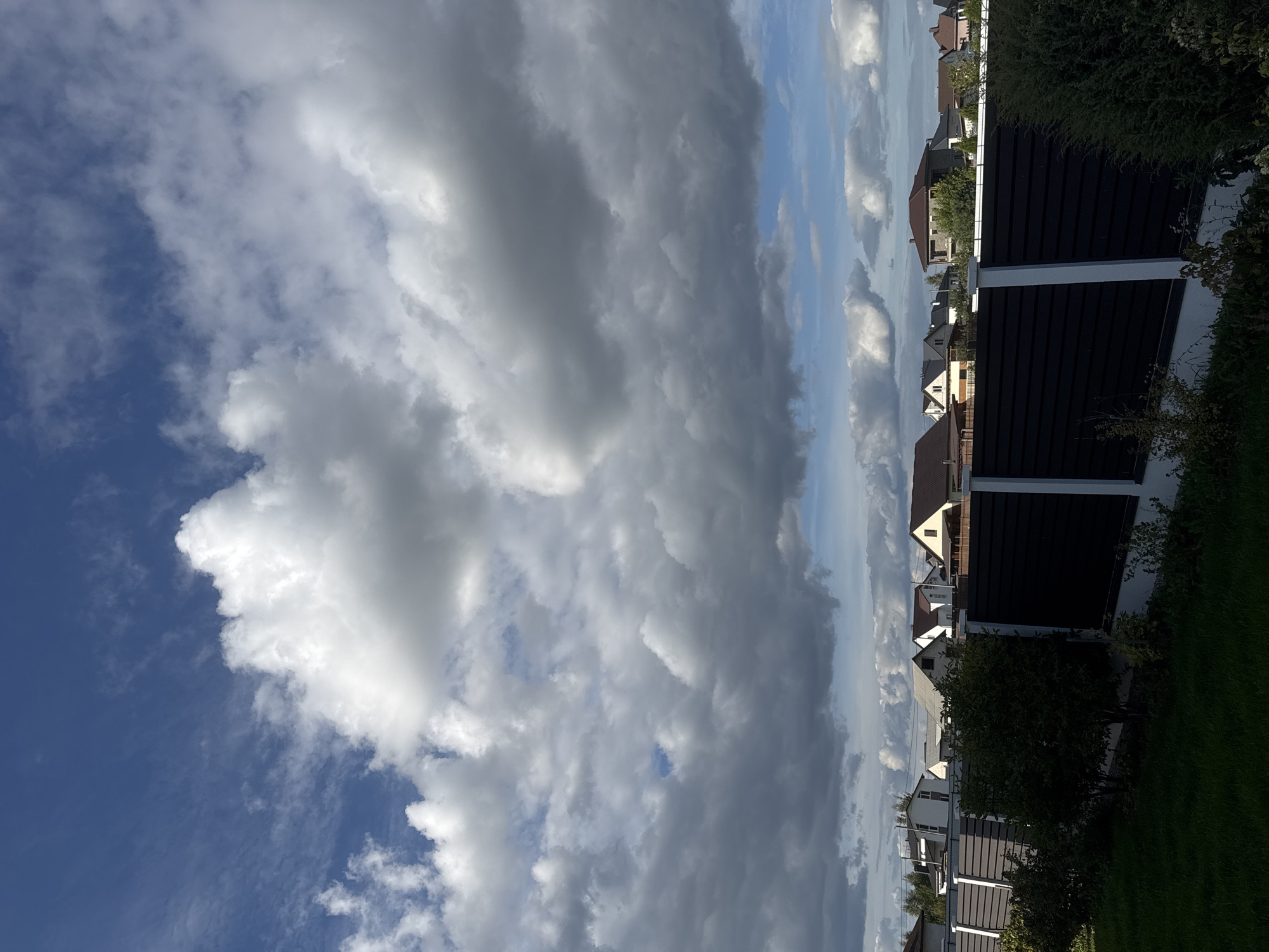 The image depicts a suburban landscape under a dynamic sky, likely taken in the Бараўлянскі сельскі Савет, Belarus. The foreground features a neatly manicured lawn and a dark, horizontal slatted fence with white vertical posts. Beyond the fence, a row of residential houses with pitched roofs in various colors and styles are visible. Lush green trees and bushes are interspersed among the houses and along the fence line. The sky above is dramatic, filled with a mix of bright white cumulus clouds and darker, more ominous storm clouds, with patches of clear blue sky peeking through. The overall impression is one of a residential area on a day with changing weather conditions.