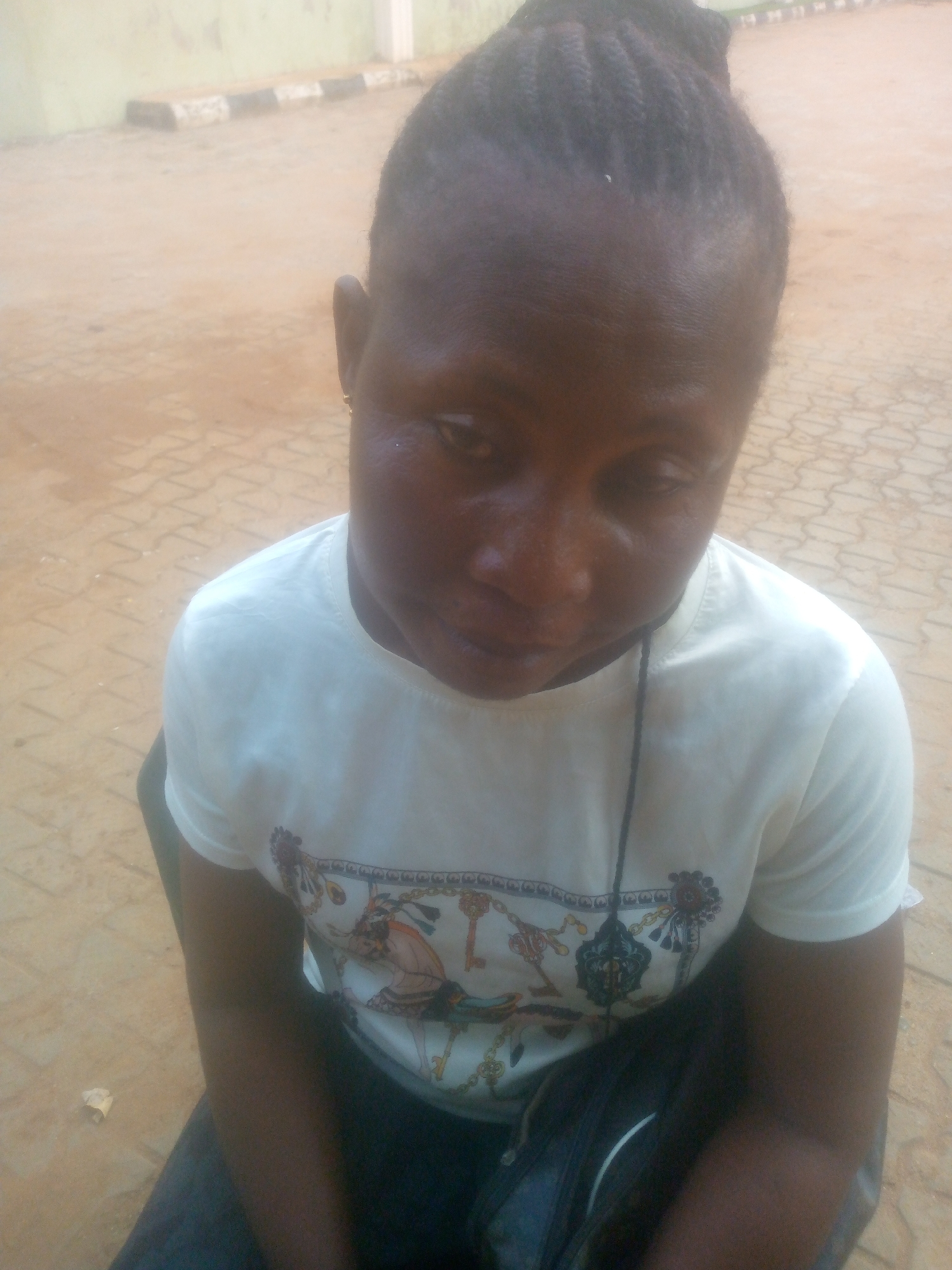 A high-angle, medium-shot photograph features an individual with dark skin and braided hair, looking downwards and slightly to their right. They are wearing a white, short-sleeved t-shirt with a complex, colorful pattern across the chest, which includes what appear to be golden chains, various keys, and illustrative figures or crests. A thin, dark string or cord hangs vertically in front of their shirt. A small golden earring is visible in their right ear.
The background consists of light brown or tan ground, paved with geometric interlocking tiles. Further back, a pale-colored wall, possibly light green or yellow, is visible, along with a low curb or barrier featuring black and white stripes. The lighting suggests it is daytime, with soft, even illumination. The person appears to be in a static pose, possibly seated or crouching, in an outdoor or semi-outdoor urban setting. A small, crumpled piece of white paper is visible on the ground near the subject's left arm in one of the provided crop images. No legible text is visible within the frame.