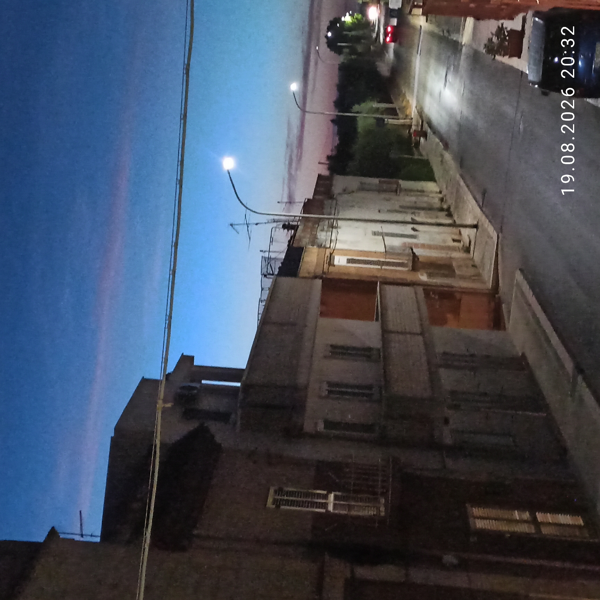 The image depicts a street scene in Bitonto, Italy, at dusk. The sky is a deep blue transitioning to a lighter hue with a faint pinkish-orange streak near the horizon. Buildings line the left side of the street, with a few windows illuminated. The street itself is paved and appears to be wet, reflecting the light from streetlamps. Two streetlights are visible, one on the left and another further down the street on the right, casting pools of light. A red car is driving away from the camera, its taillights visible. The image also features a timestamp in the bottom right corner: "19.08.2026 20:32". No people are clearly visible in the scene. The overall atmosphere is calm and quiet, characteristic of a residential street at the end of the day.