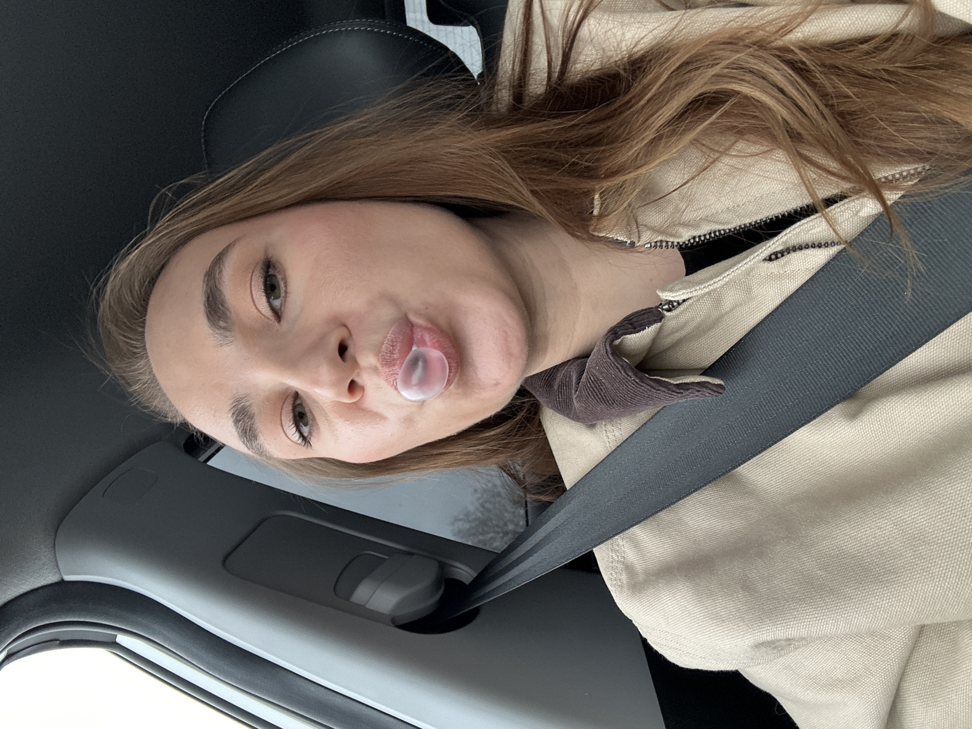 A young woman is seated in a car, wearing a light brown jacket and a seatbelt. She is blowing a bubble with gum. The interior of the car is visible, including the window, door panel, and black seats. The location of the scene is not identifiable from the image.