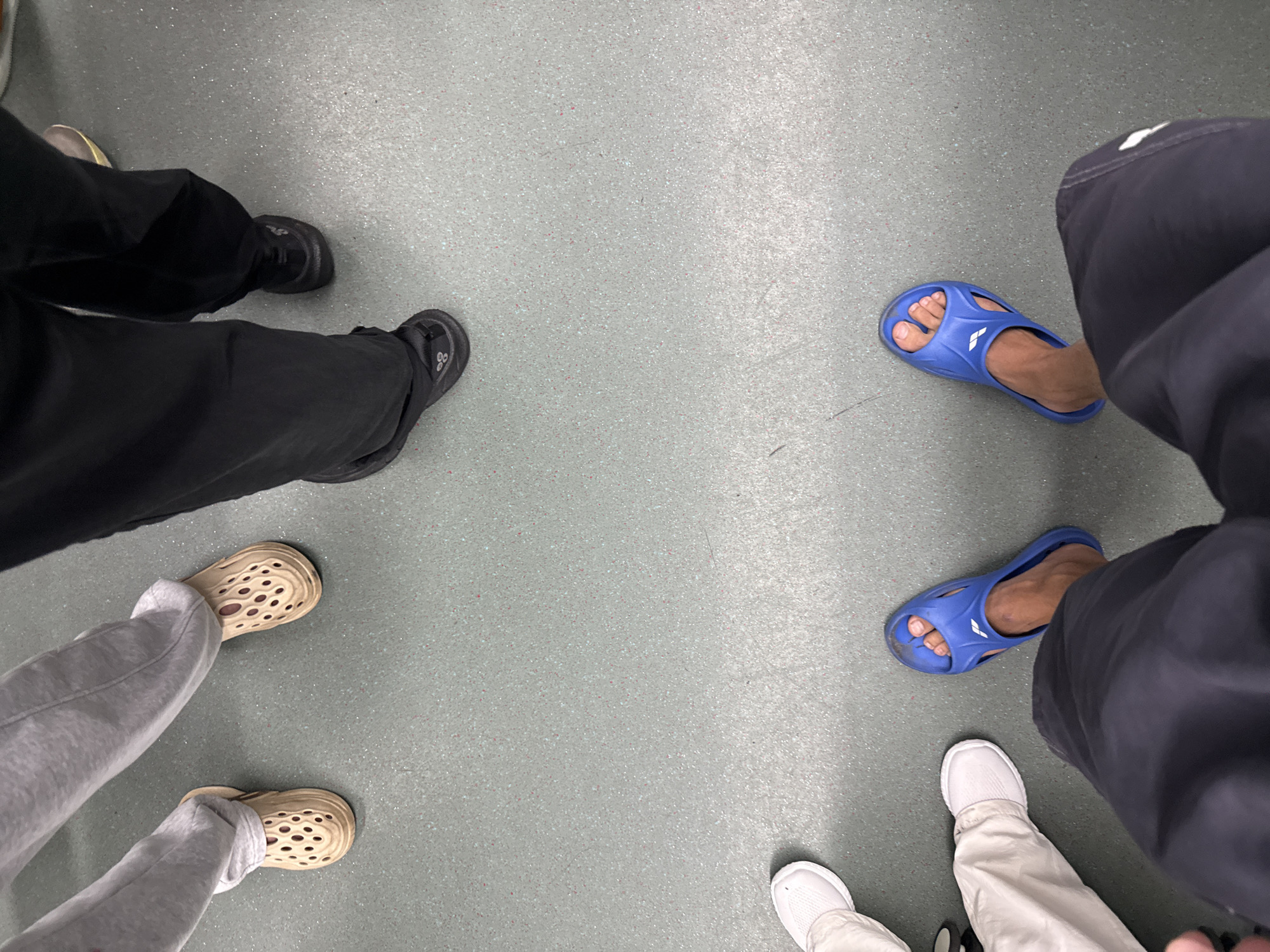 The image shows the legs and feet of at least four individuals standing on a speckled grey floor, possibly in a public transit area in Haizhu District, China. From top left to bottom right, the visible footwear includes: a pair of tan, perforated clogs; black athletic shoes; white sneakers; and a pair of blue slides. The individuals are wearing casual clothing, including grey sweatpants, black pants, and white pants. The scene is viewed from a high angle, looking down at the feet.