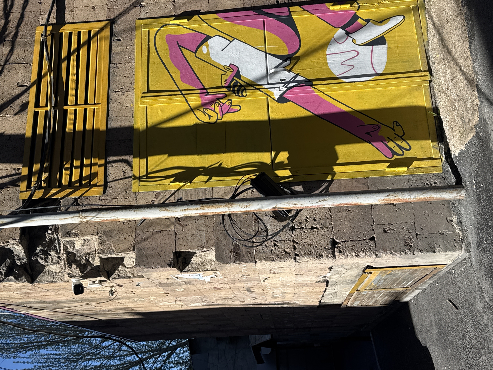 The image depicts a section of a Yerevan, Armenia city street viewed from below. Above, an old brick wall supports a metal pipe running horizontally. Two yellow painted metal doors are set into the wall: one with vertical bars, the other with an image of a person in white, pink, and black with the letter "M." Shadow patterns cast on the yellow doors suggest late afternoon sunlight. Below, a sidewalk of worn concrete tiles and part of a street are visible, obscured by the angle. A security camera is mounted under the sidewalk. In the lower left, the reflection of trees and a blue sky is visible on a parked car's surface.