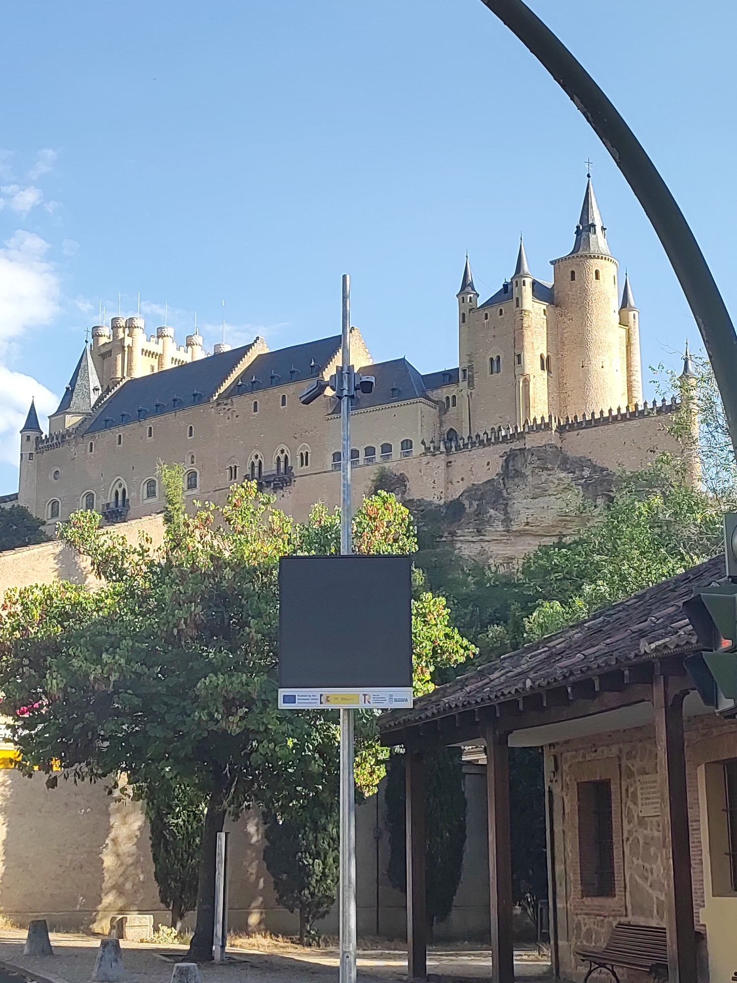 The image depicts the Alcázar of Segovia, a historic royal palace and fortress located in Segovia, Spain. The Alcázar is situated on a rocky promontory overlooking the confluence of two rivers, with its distinctive ship-like shape and numerous spires and turrets dominating the skyline. The fortress is constructed of stone and appears well-preserved, with its crenellated battlements and tall, conical towers visible.
In the foreground, there is a modern digital display board mounted on a pole, with some signage at its base. To the right of the display, a section of a tiled roof structure is visible, appearing to be a small building or covered area. Green trees with lush foliage are scattered throughout the scene, some in the mid-ground behind the display board and others in the background, partially obscuring the lower sections of the Alcázar.
The sky is a clear blue with a few wispy clouds. The overall lighting suggests it is daytime, likely morning or afternoon, with sunlight illuminating the castle and casting shadows. There are no people clearly visible in the image, and the scene appears to be a public space, possibly a viewpoint or tourist area. The image captures a grand architectural landmark set against a natural and urban backdrop.