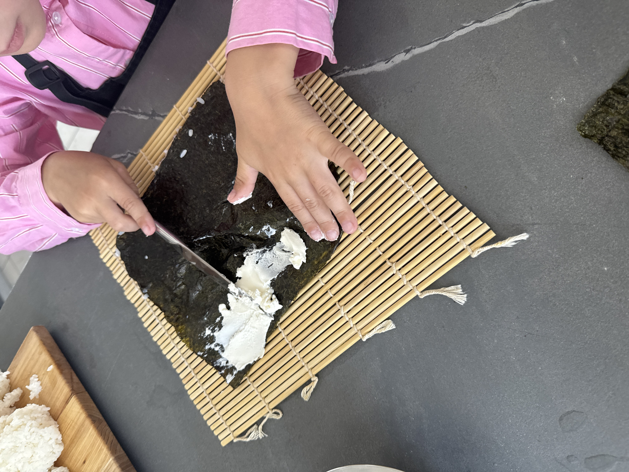 A child, wearing a pink and white striped shirt and a black apron, is preparing sushi on a bamboo rolling mat. The child is spreading a white creamy substance, likely cream cheese or mayonnaise, onto a sheet of nori seaweed with a knife. Several grains of rice are visible on the nori. To the left of the mat, on a wooden cutting board, is a pile of cooked white rice. The surface beneath the mat is a dark grey countertop, possibly slate, with a faint white veining. The scene is indoors, likely in a kitchen. This image does not provide any specific details that would allow for localization to Izhevsk, Russia; it is a generic kitchen scene.