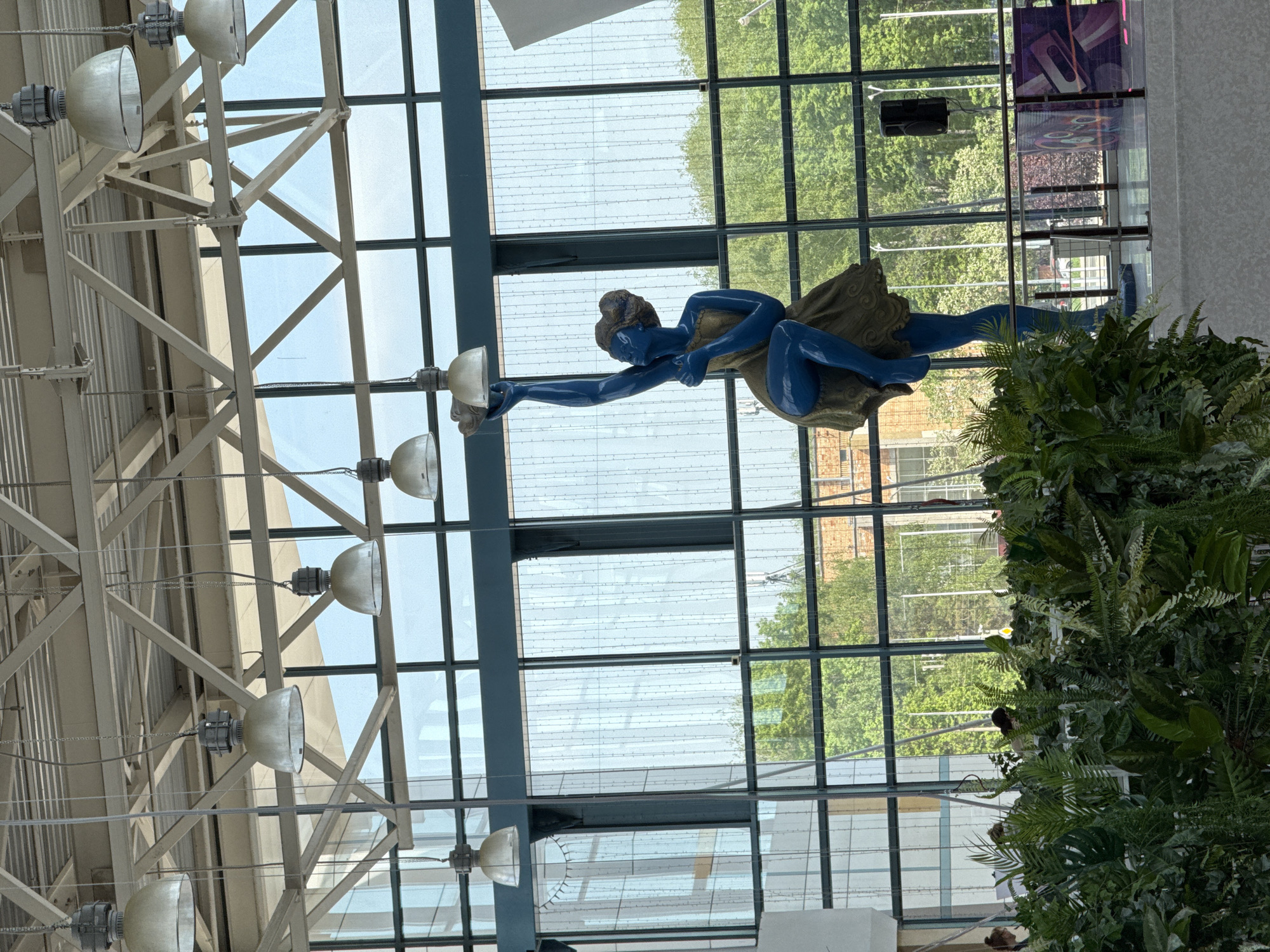 The image shows an indoor view in Nizhny Novgorod, Russia. A blue statue of a reclining figure holding a lamp is suspended in front of a large glass window. Outside the window are trees and buildings. The interior features an exposed ceiling with metal beams and lighting fixtures. A wall of green foliage is visible on the right.