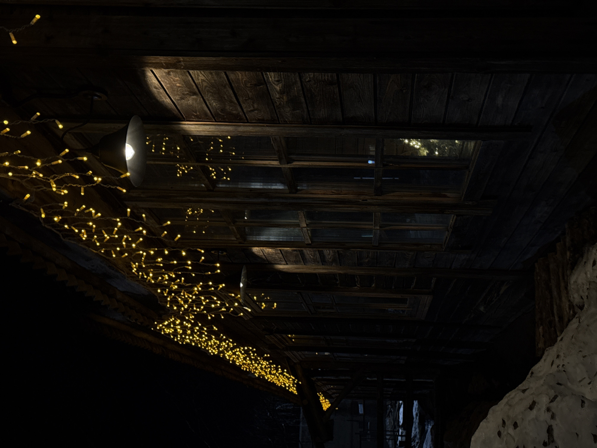 The image captures a dark, low-lit interior space, possibly a wooden structure or building. The ceiling is composed of dark wooden planks, and a series of rectangular openings, resembling windows or skylights, are visible. Strings of small, warm-toned lights are draped across the beams and under the eaves, providing the primary source of illumination. A lamp with a conical shade directs light downward. To the right, a textured surface that could be snow or a light-colored wall adds contrast to the scene. The city and its exact location is unknown.