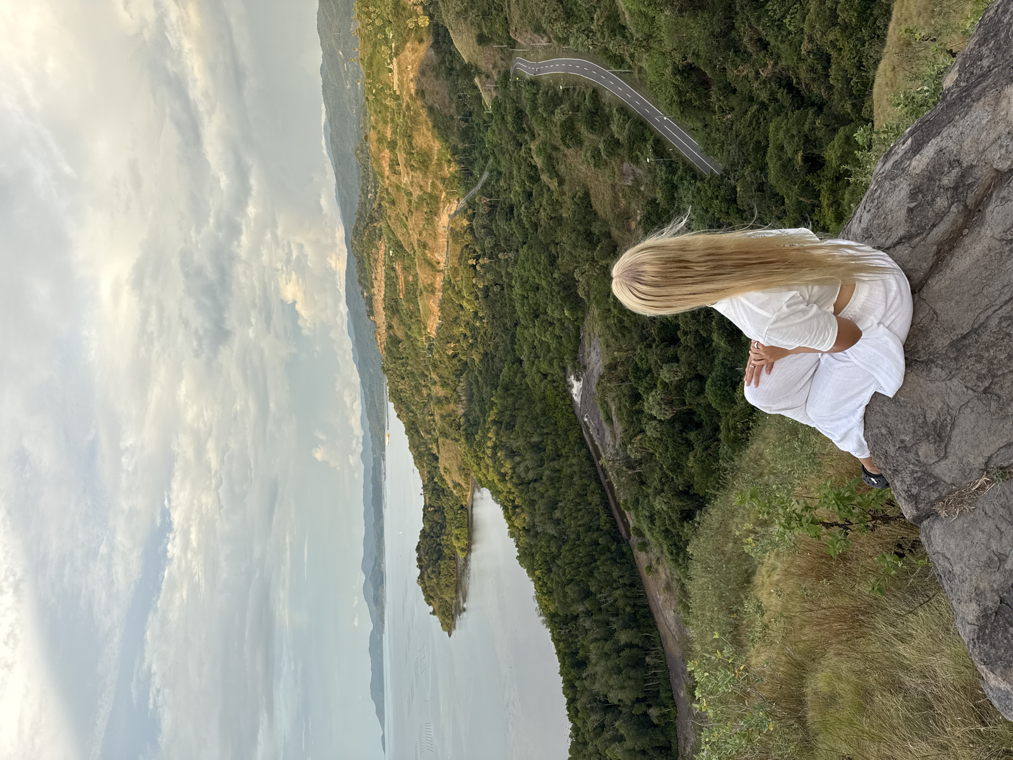 A woman with long blonde hair sits on a rocky precipice overlooking a scenic landscape in Labuan Bajo, Indonesia. She is dressed in white clothing, and her posture suggests contemplation. Below her, a lush green forest extends towards a body of water that appears to be a bay or a lake. A winding road is visible through the forest. The sky above is partially covered with clouds. The photograph is taken from a high angle, emphasizing the height and the scale of the surroundings.