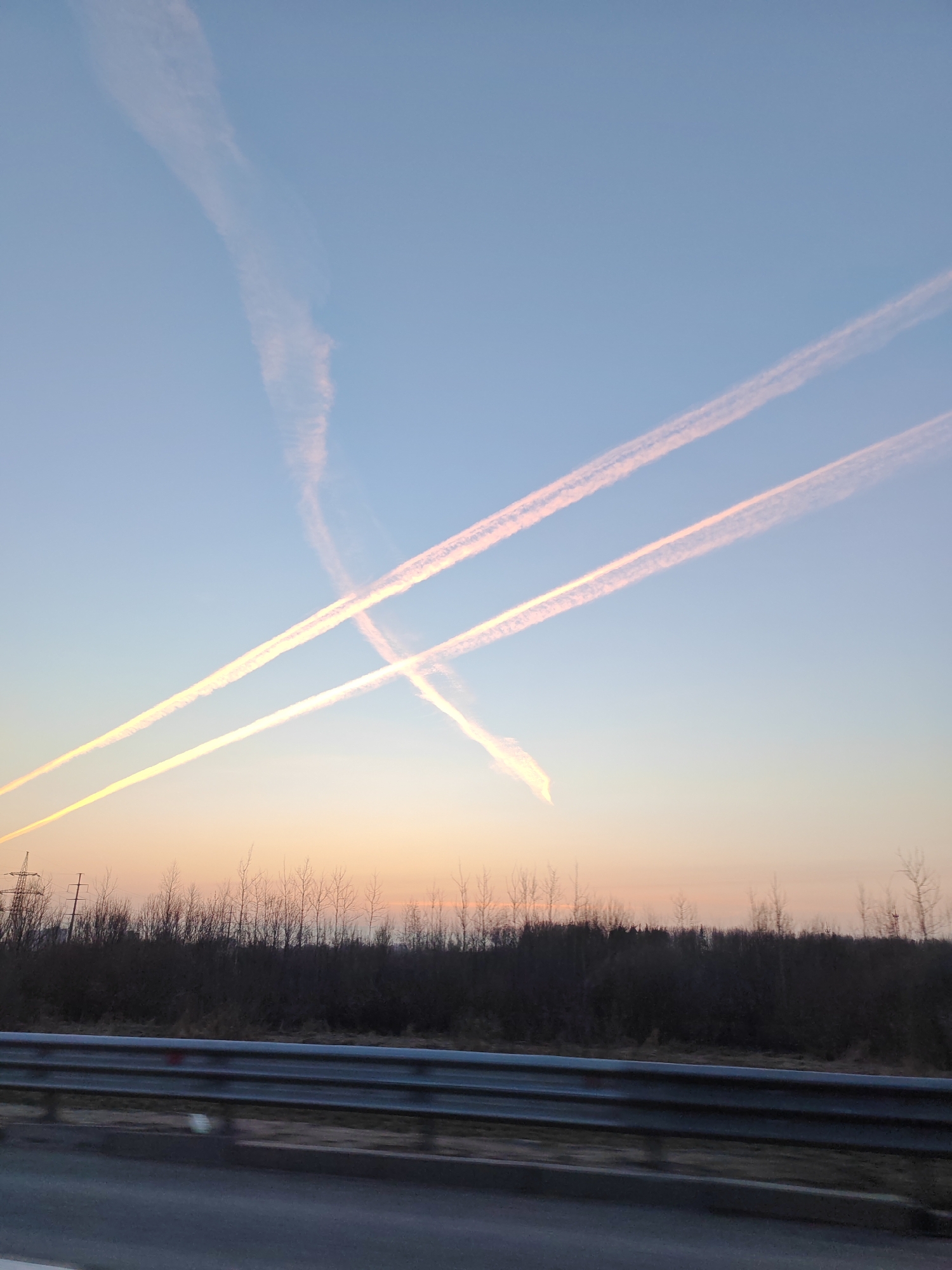 The image shows a view from a roadside in Zanevka, Russia. The foreground features a gray road surface and a metal guardrail. Behind the guardrail, there's a dark line of bare trees or bushes against a sky transitioning from a pale orange or pink near the horizon to light blue above. Vapor trails from aircraft crisscross the sky, creating an X shape in the center and another trail higher up. Electrical power lines are visible in the lower left corner. No people or other subjects are identifiable in the image. The time appears to be around sunset or sunrise.