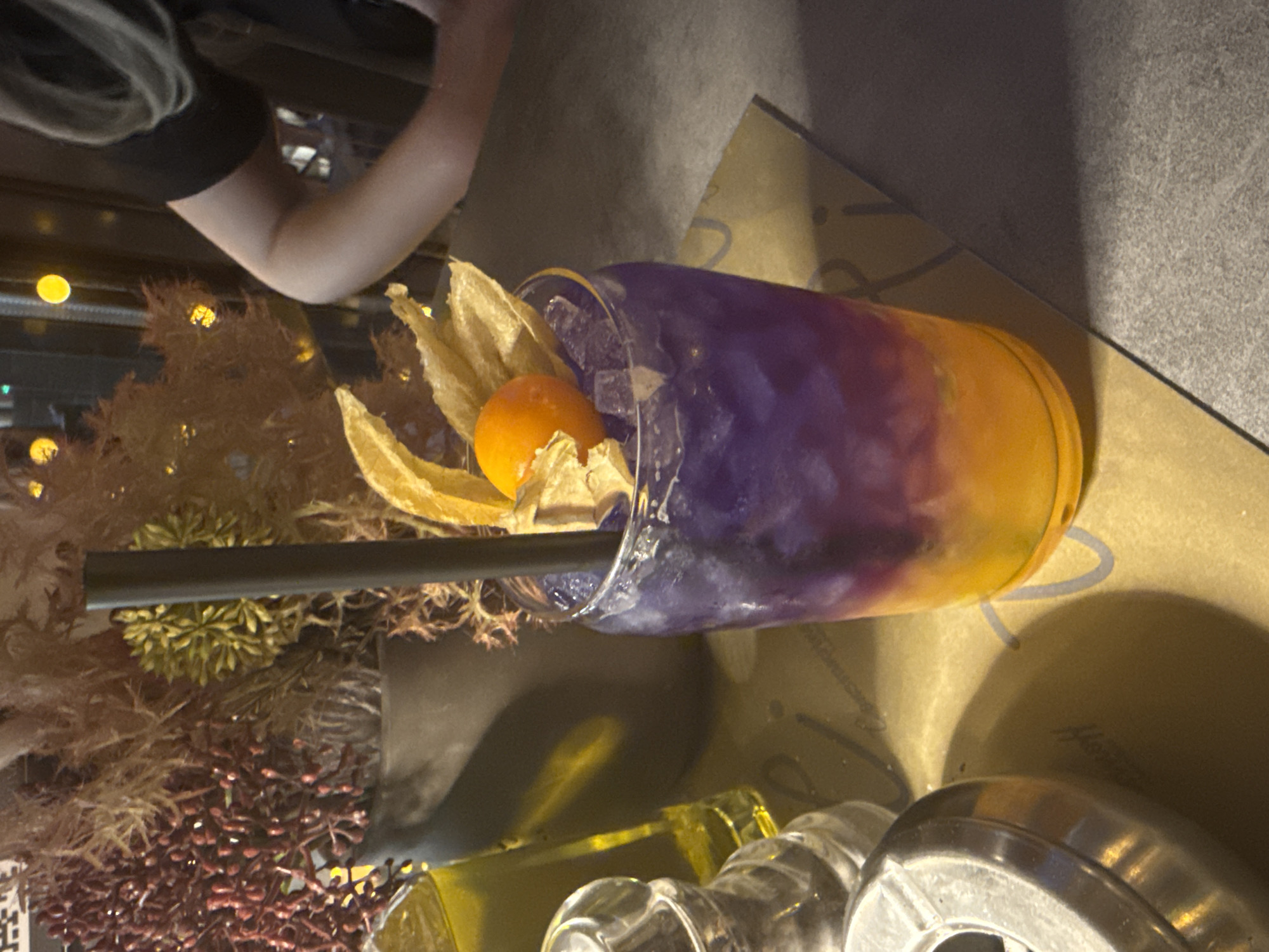 A colorful cocktail in a plastic cup sits on a table next to a floral arrangement. The drink is layered with purple and orange hues and garnished with a physalis. A black straw is inserted into the drink. In the upper left corner of the frame, part of a person's arm and shoulder are visible. The table is likely in a bar or restaurant in the city of Sofia, Bulgaria.