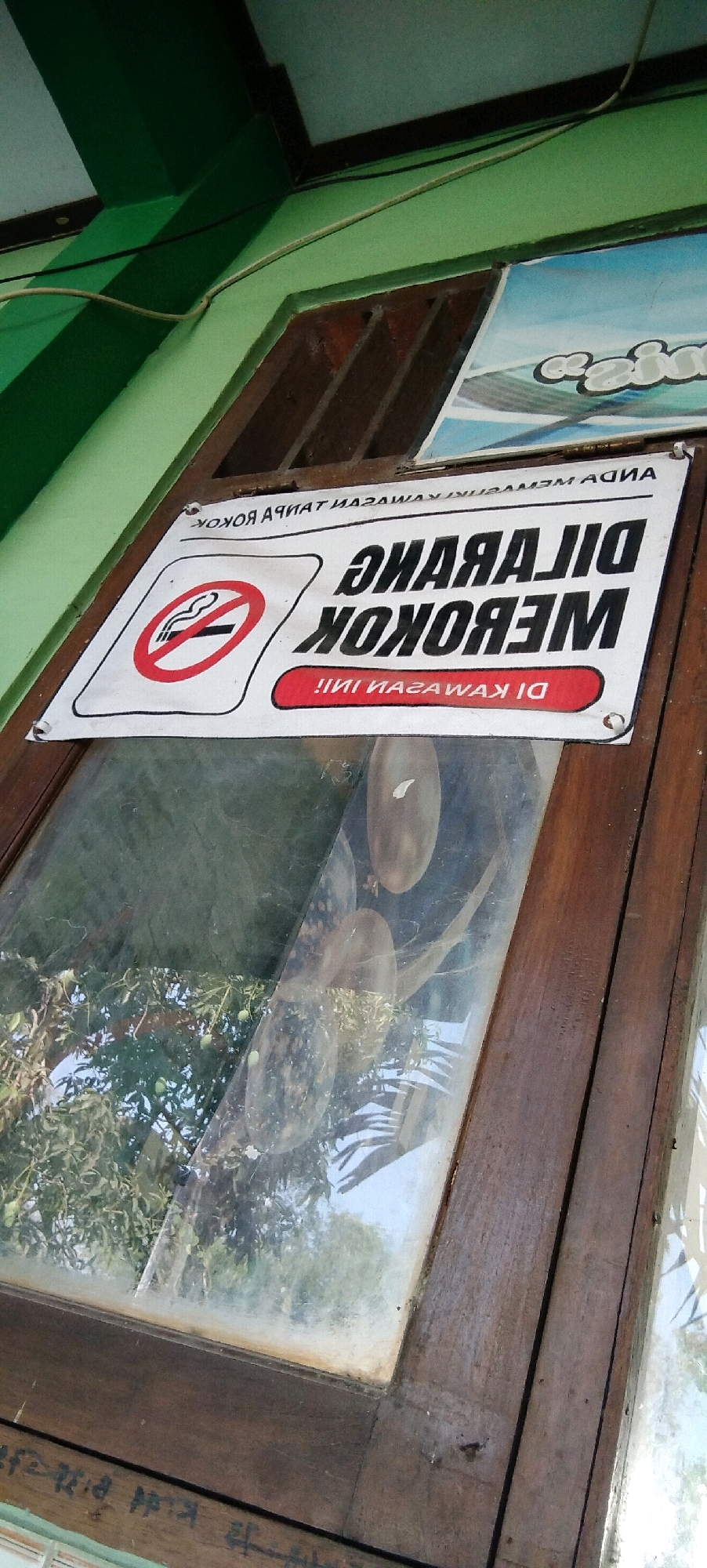 The image shows a "NO SMOKING" sign attached to a wooden framed window. The sign is white with black text and a red circle with a diagonal line through a cigarette. The text on the sign is in Indonesian and reads "DILARANG MEROKOK DI KAWASAN INI" which translates to "SMOKING IS PROHIBITED IN THIS AREA". The window is slightly grimy and reflects the outdoors, showing trees and greenery. The sign appears to be located in a building in Wadang, Indonesia, as indicated by the provided context. The overall scene depicts a common public health notice in a tropical setting.