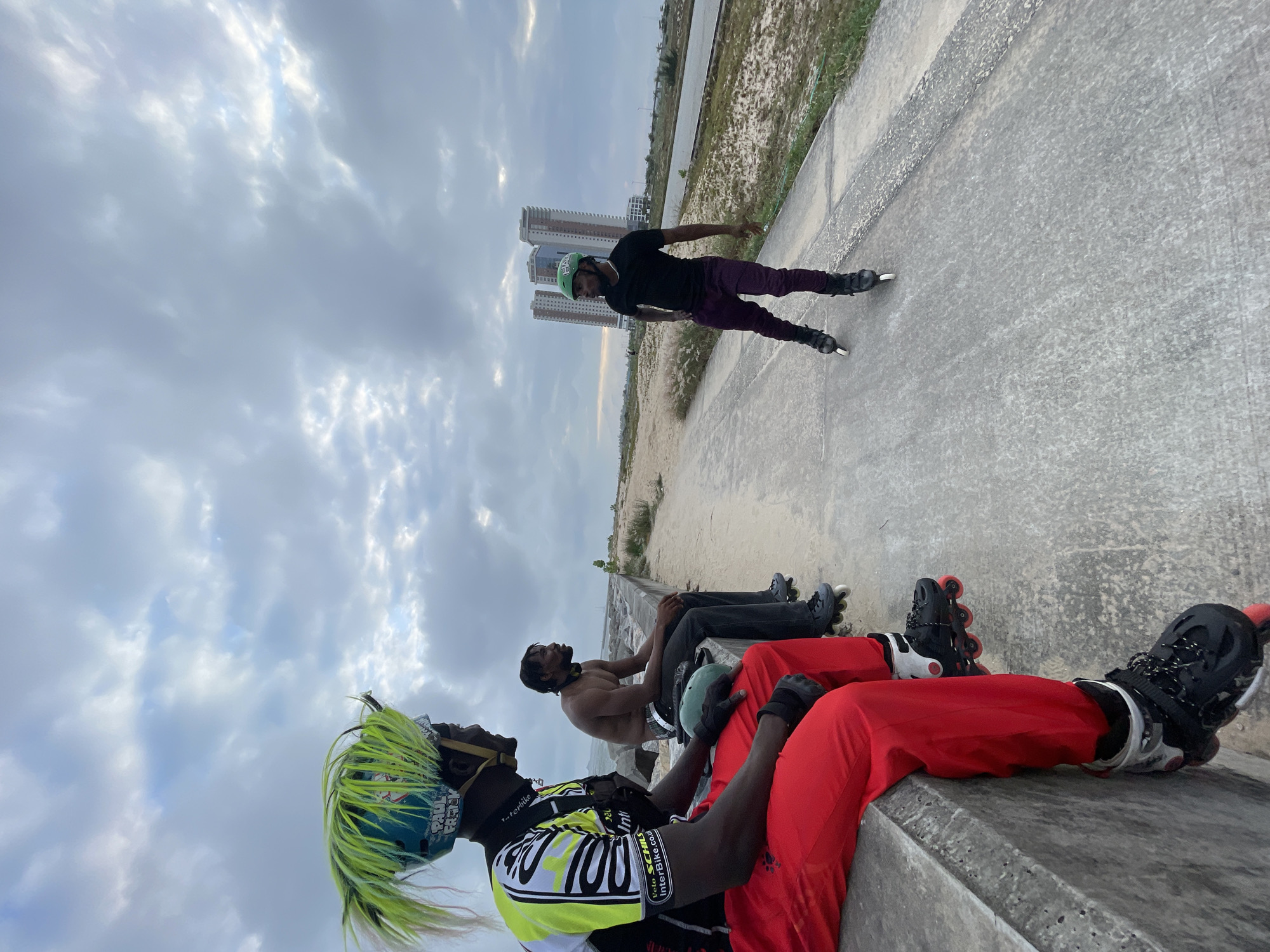 In Ilado, Nigeria, three individuals are engaged in rollerblading. Two men are seated on a concrete barrier, facing away from the camera. The man on the left wears a helmet with bright green, spiky hair, a black and yellow cycling jersey, and red pants with rollerblades. The man on the right, shirtless, wears dark pants and rollerblades, with a green helmet beside him. A third man is rollerblading on a concrete path in the background, wearing a black t-shirt, dark pants, and a green helmet. In the distance, modern high-rise buildings and a body of water are visible under a cloudy sky. The environment appears to be a coastal area with sparse vegetation and sandy ground.