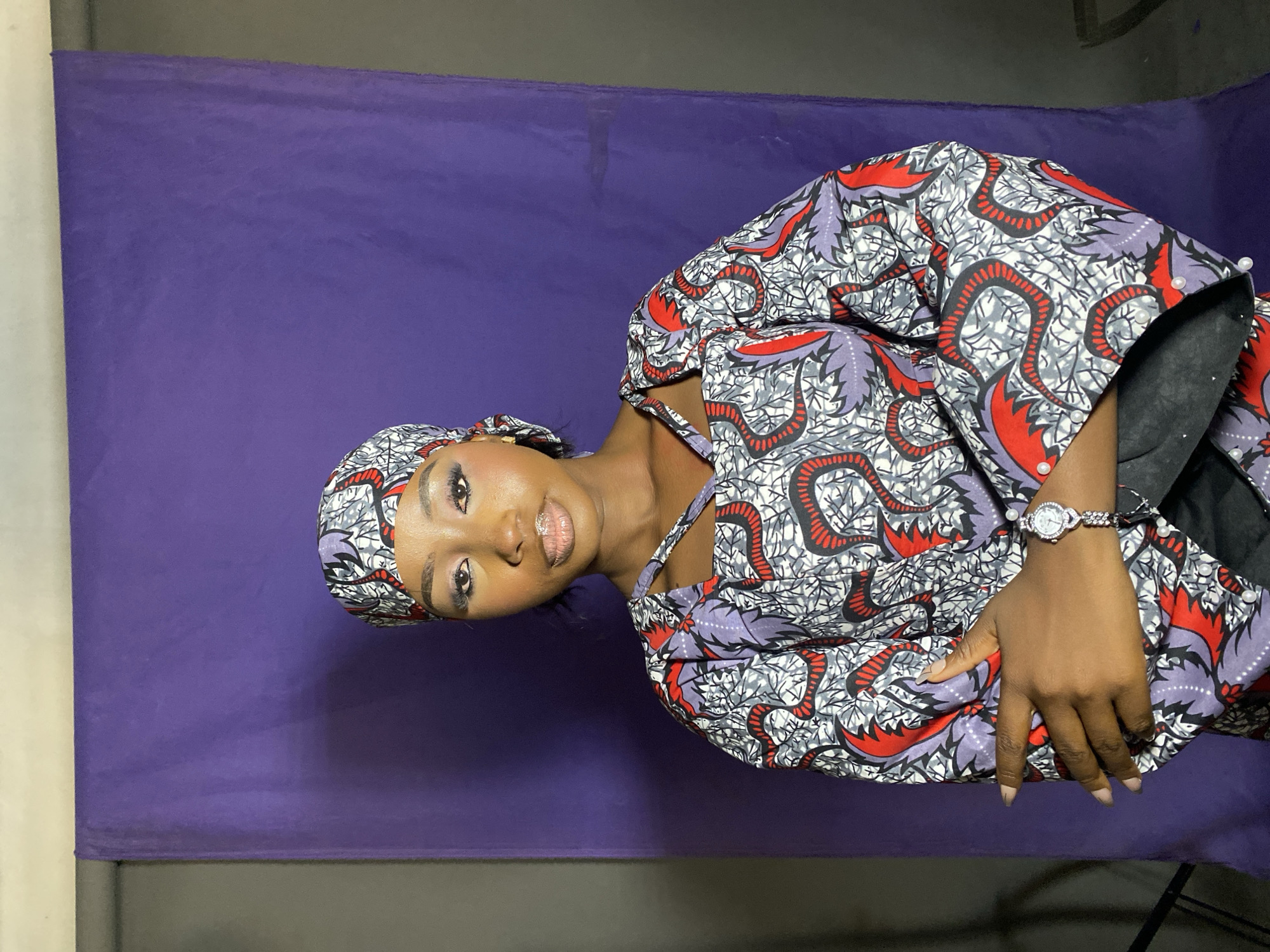 A young woman wearing a printed headwrap and robe is photographed against a purple backdrop. The setting seems to be indoors. She is wearing a wristwatch on her left wrist and posing with her hands together. The location is Gura Top, Nigeria.